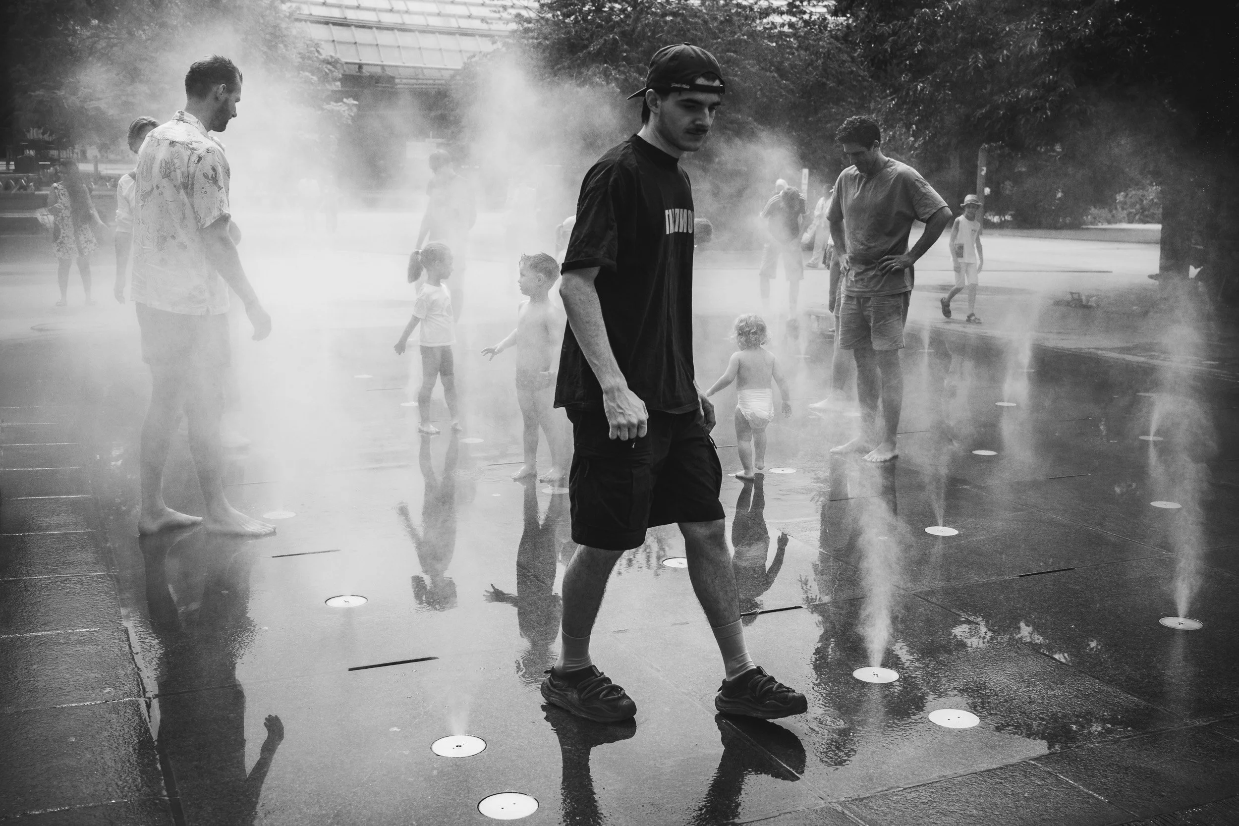People, including children, playing and walking on a wet surface with water spray or mist, outdoors in a park or public area, during the daytime.