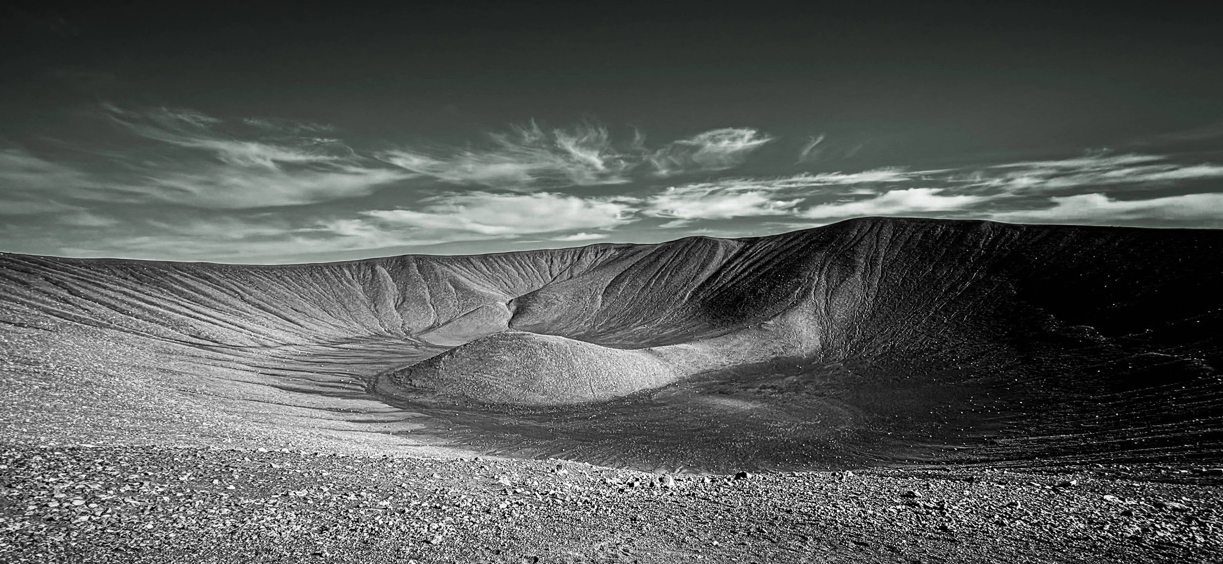 Black-and-white volcanic crater reveals sweeping ridges beneath a dramatic clouded sky.