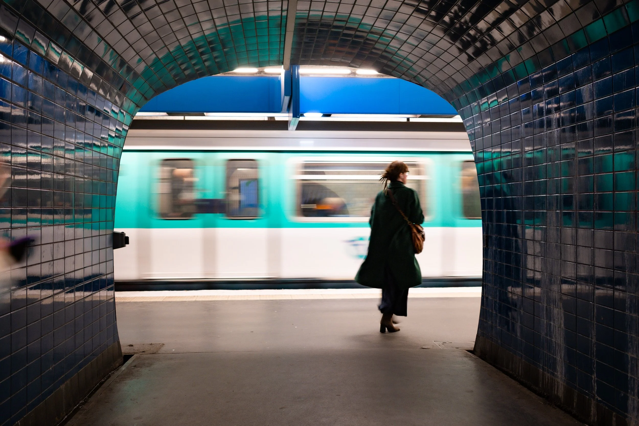 Woman walks through a blue-tiled archway as a turquoise metro train moves across the platform.