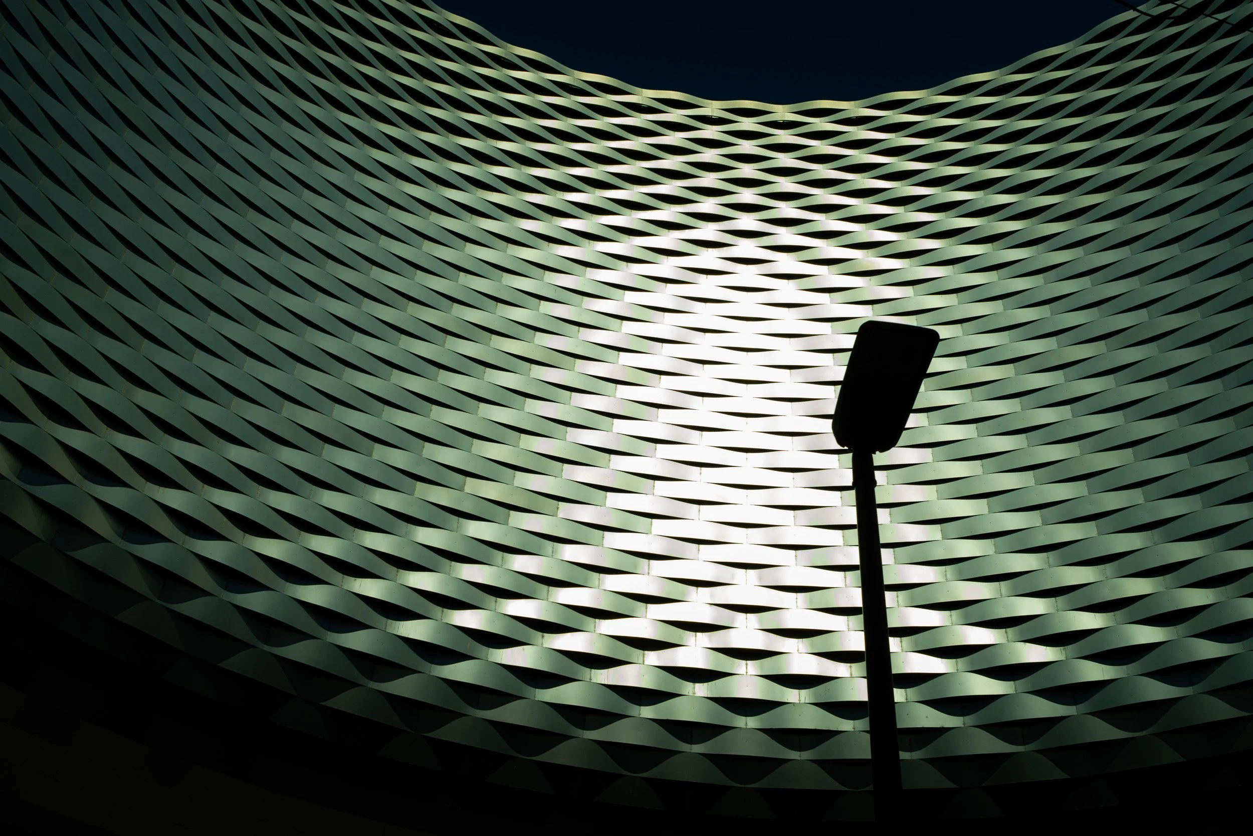 Street lamp silhouetted against an illuminated curved facade of woven metal panels.
