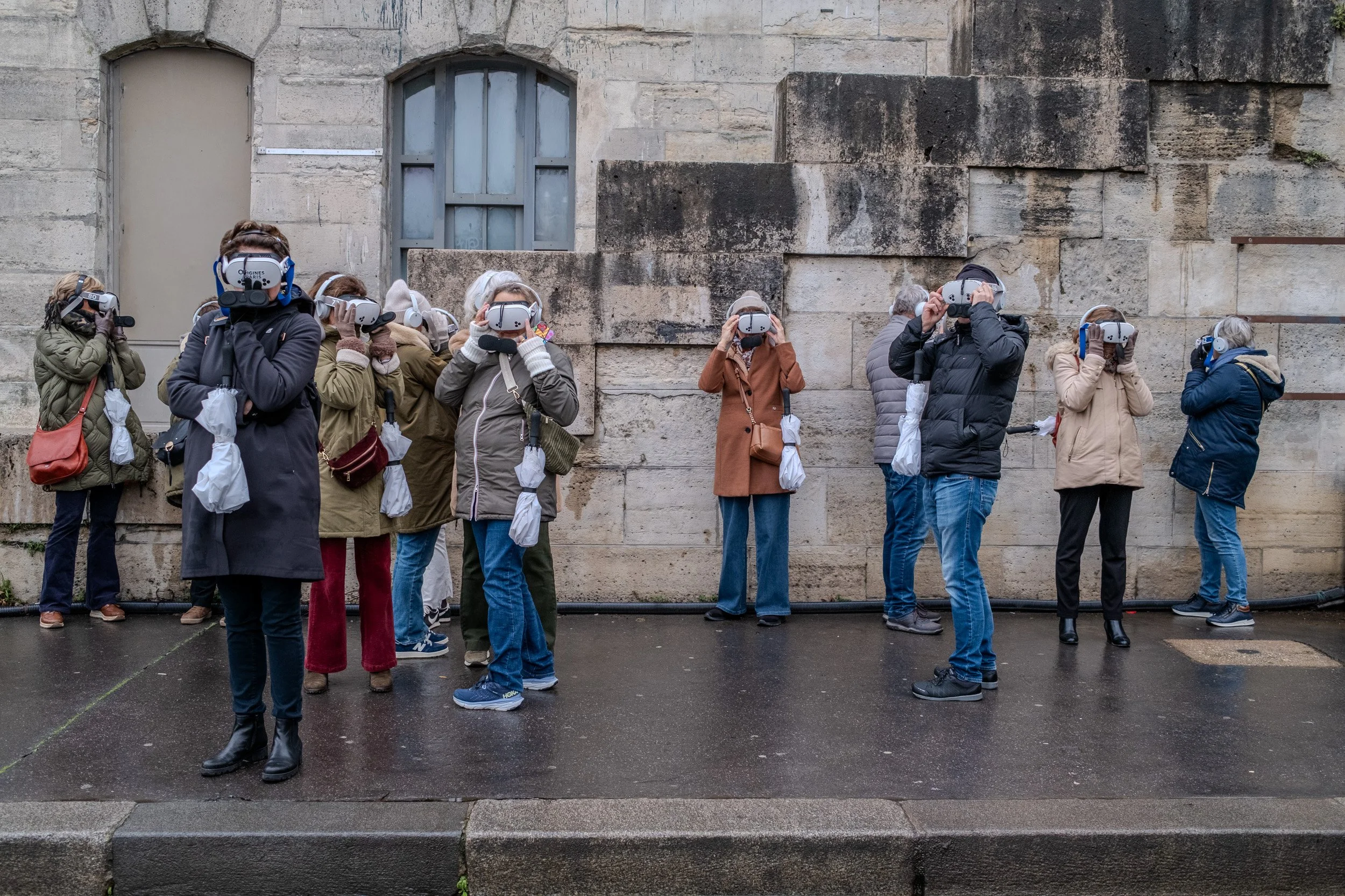 A group of adults, standing outdoors against a stone wall, wearing virtual reality headsets, possibly engaging in a virtual experience or activity.