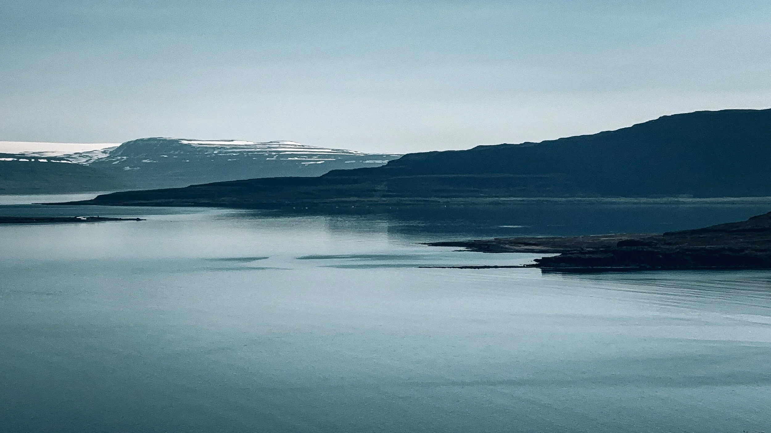 Still blue lake and layered dark hills lead toward a snow-covered plateau beneath a pale sky.