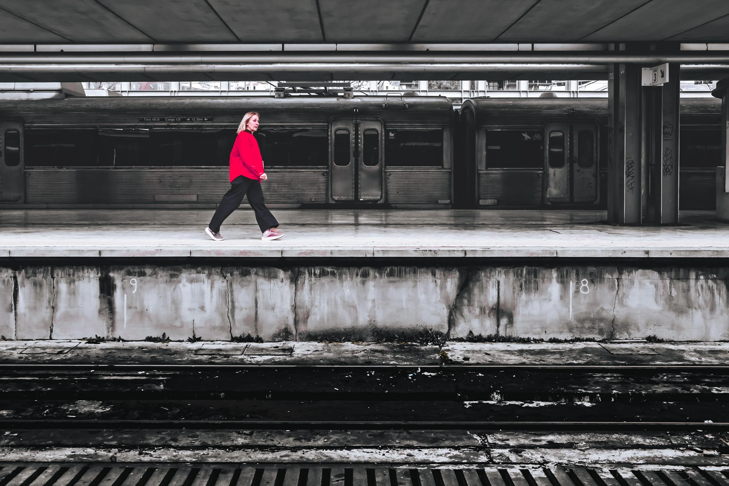 Woman in a bright red jacket walks along a railway platform in front of a dark train.