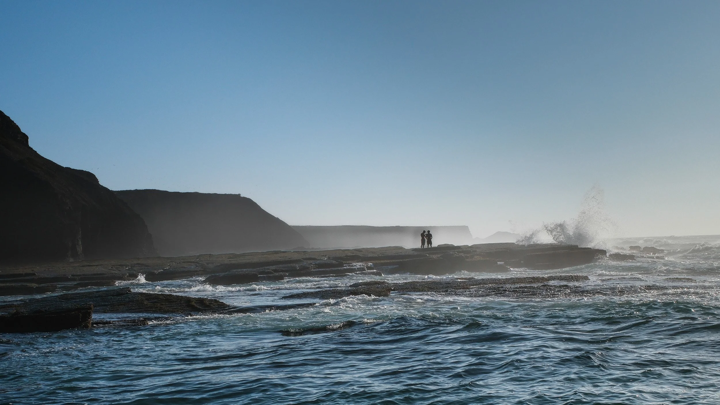 Two figures stand on a rocky coast as waves break against dark cliffs in the distance.