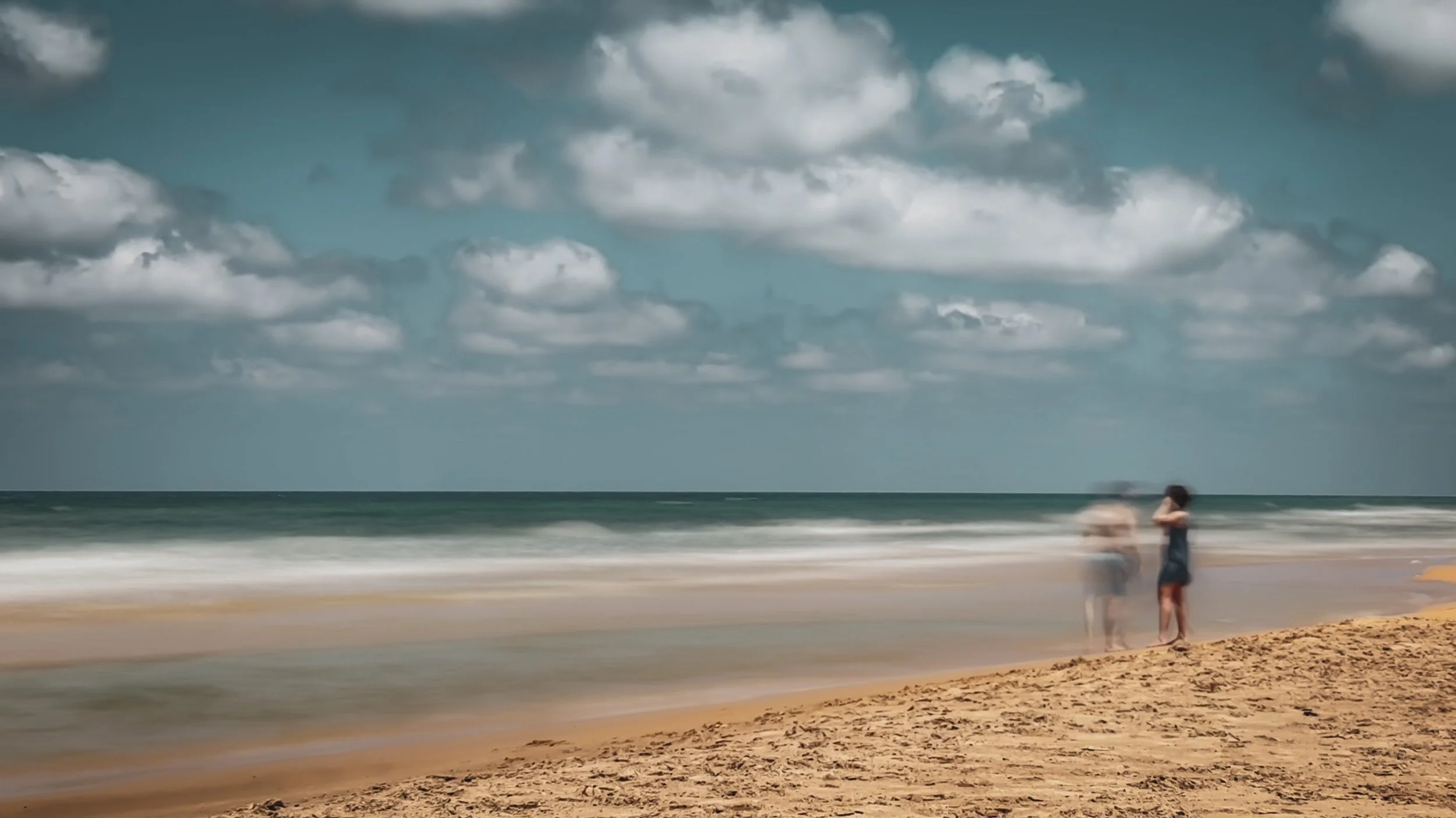 Two blurred figures stand on a sandy beach beside softly moving waves beneath scattered clouds.