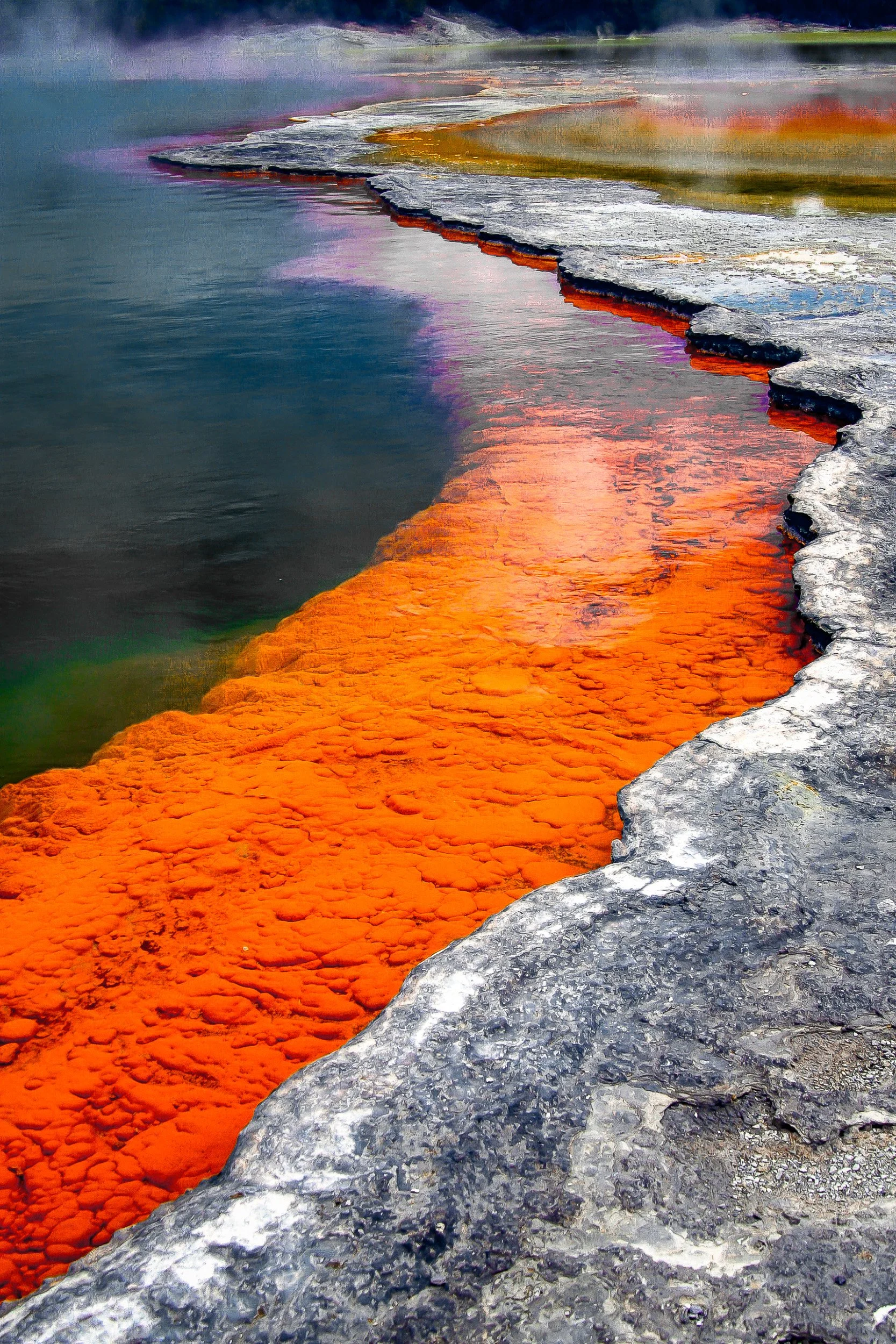 Vivid orange mineral shoreline meets dark water and pale geothermal deposits.