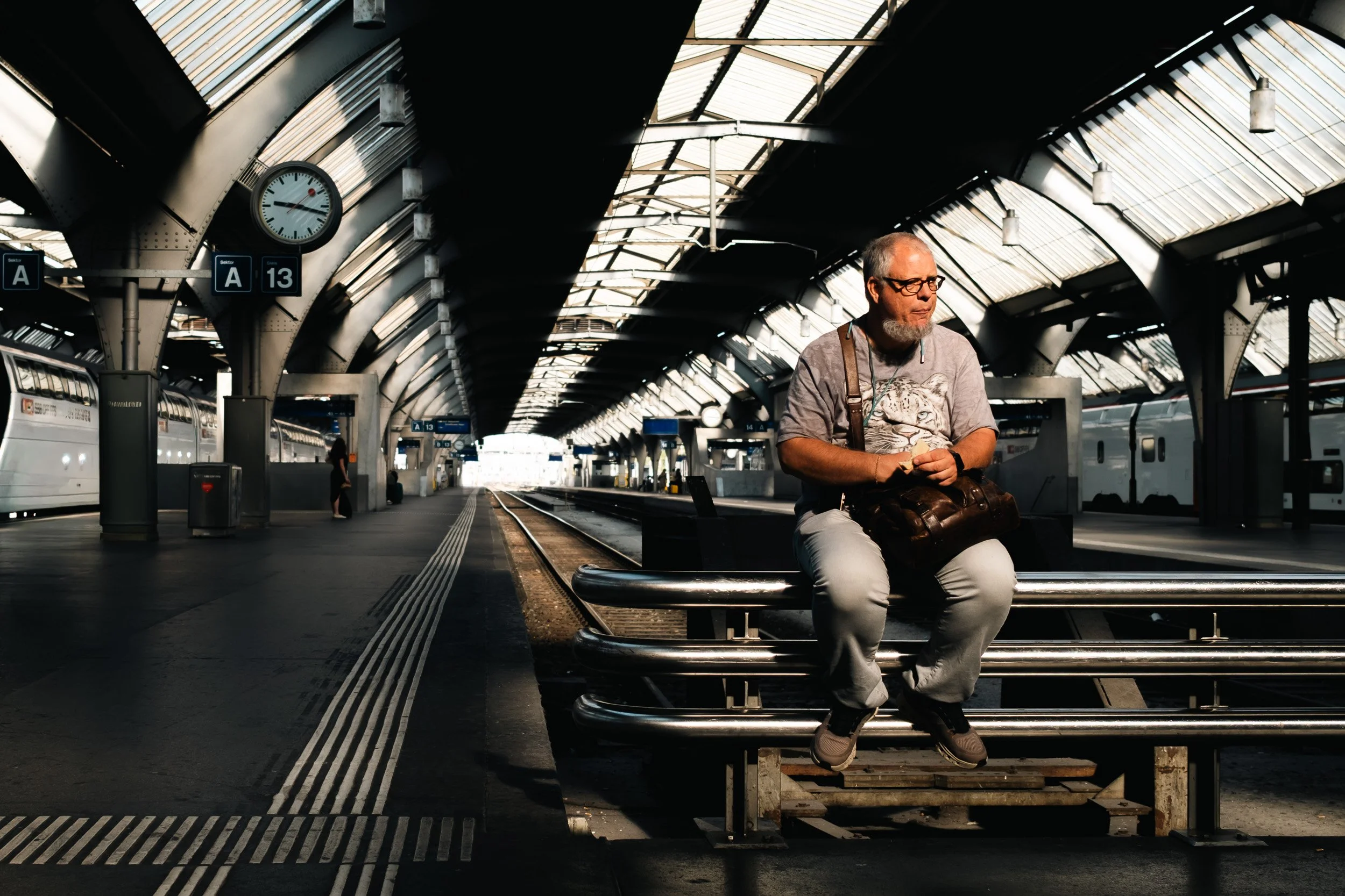 Man with a camera bag sits on a metal bench beneath a station clock and vaulted railway roof.