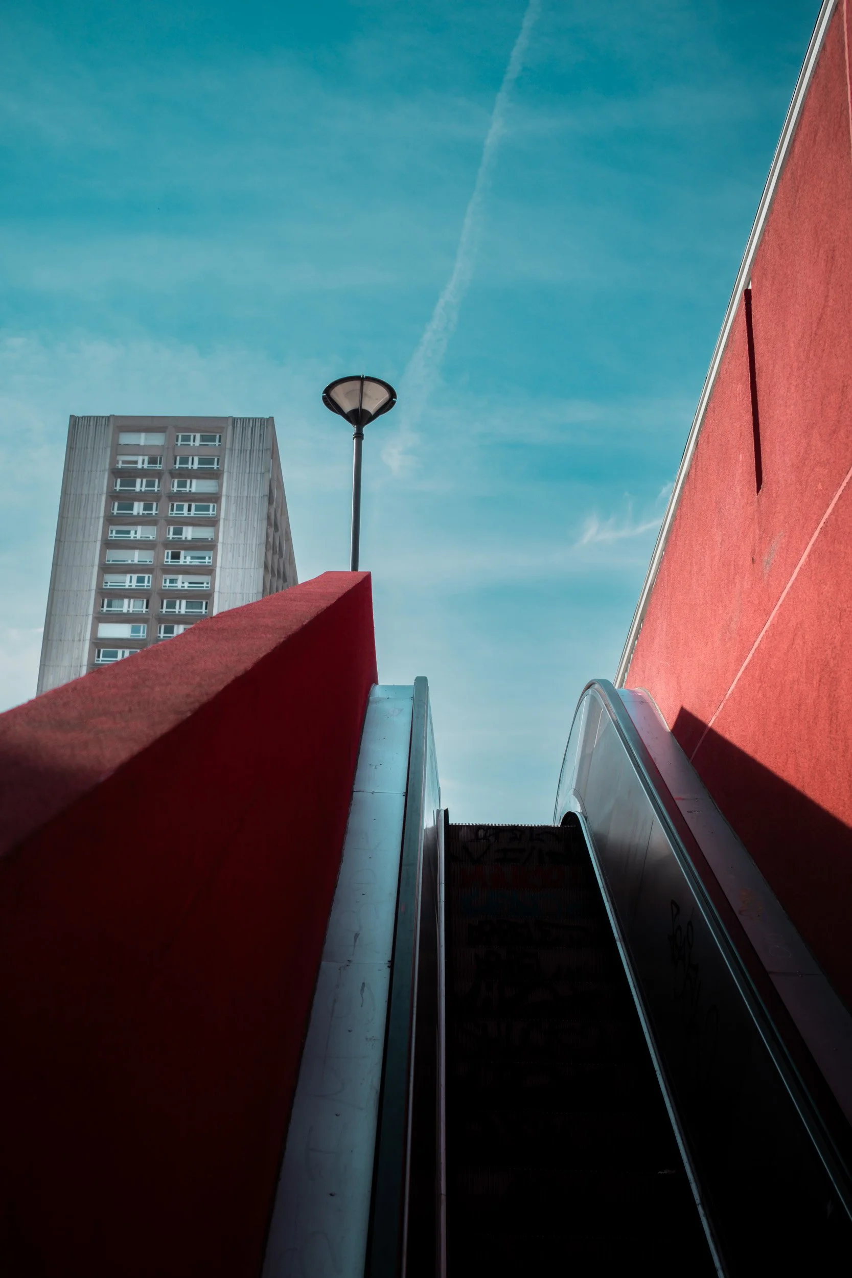 Escalator rises between red concrete walls toward a street lamp and high-rise building under a blue sky.