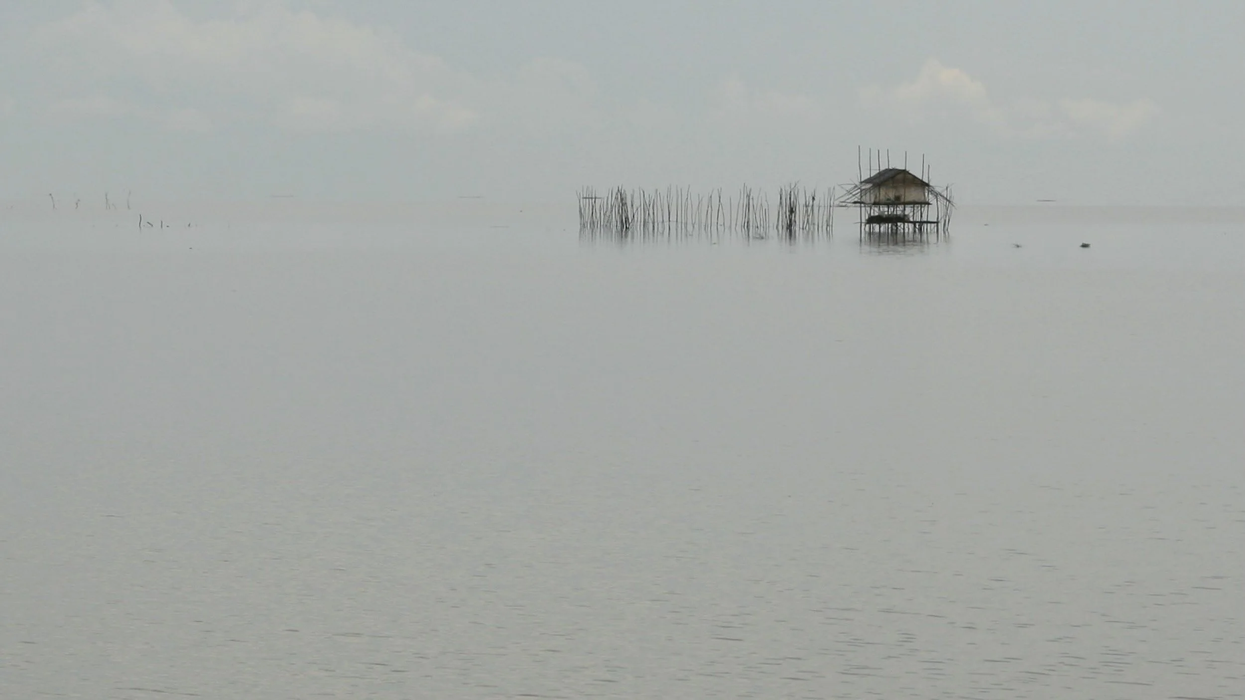 Isolated fishing hut and slender poles rise from still water in dense mist.