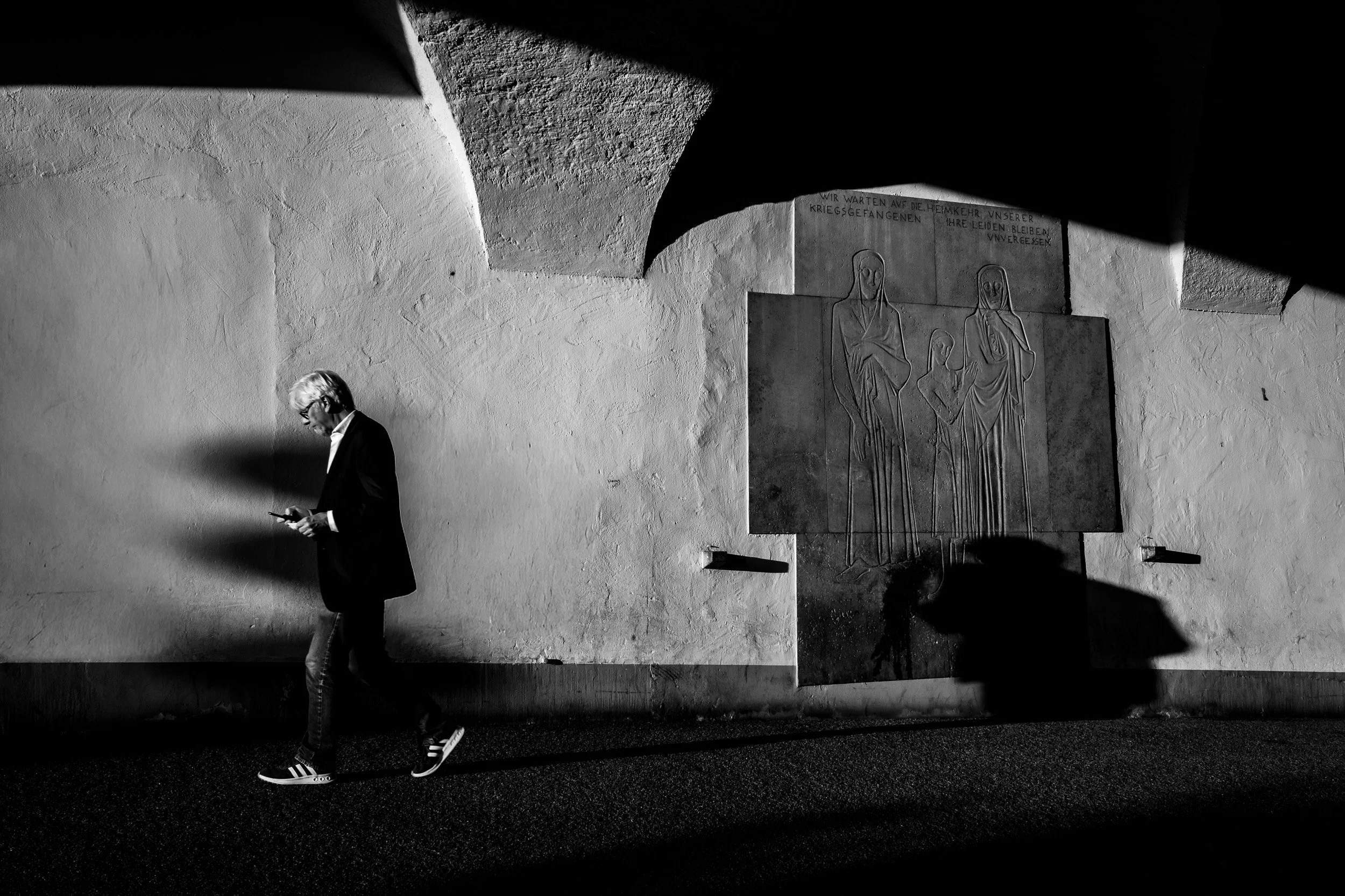 A man with blonde hair, glasses, wearing a black jacket, jeans, and sneakers, is walking while looking at his phone. The environment is a stark, textured wall with a religious sculpture or relief of three figures with a German inscription above them. The scene is in black and white with strong shadows.
