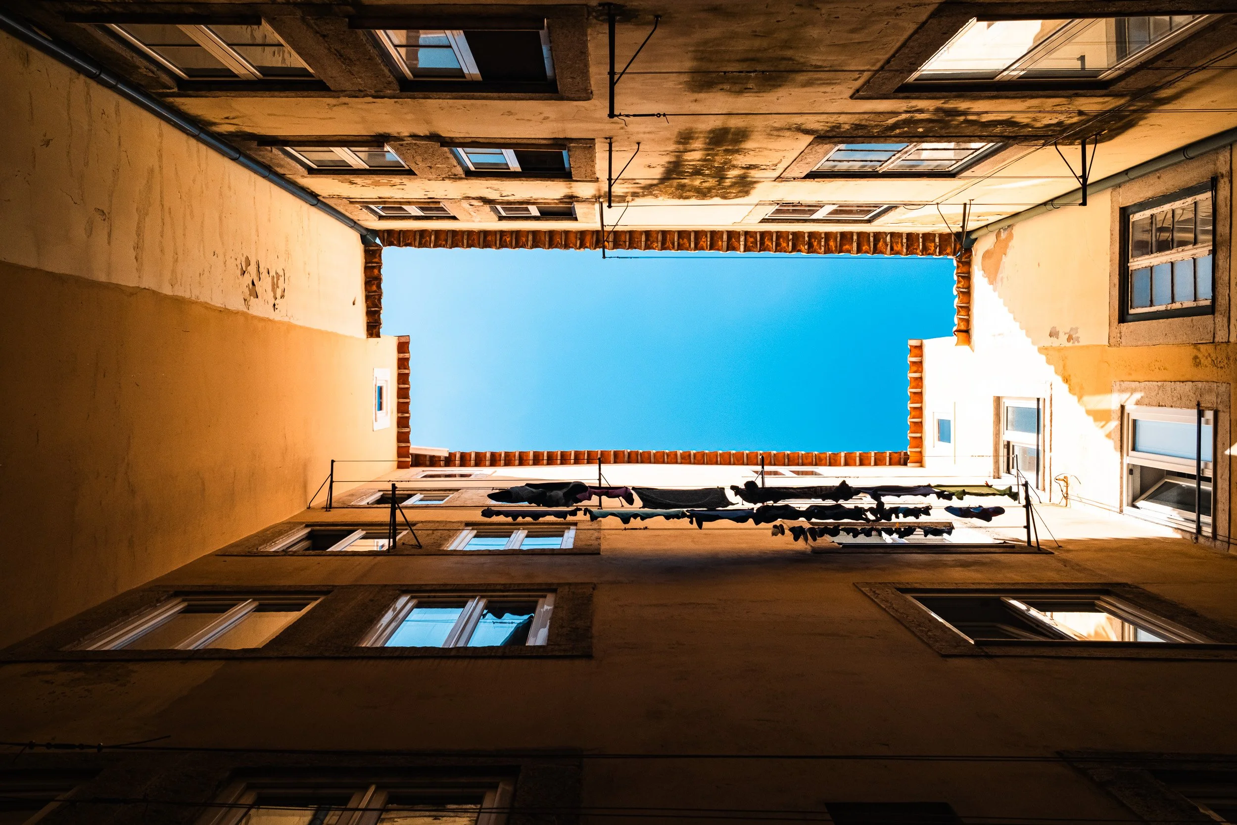 Sunlit apartment courtyard viewed upward toward a rectangle of blue sky and a line of hanging laundry.
