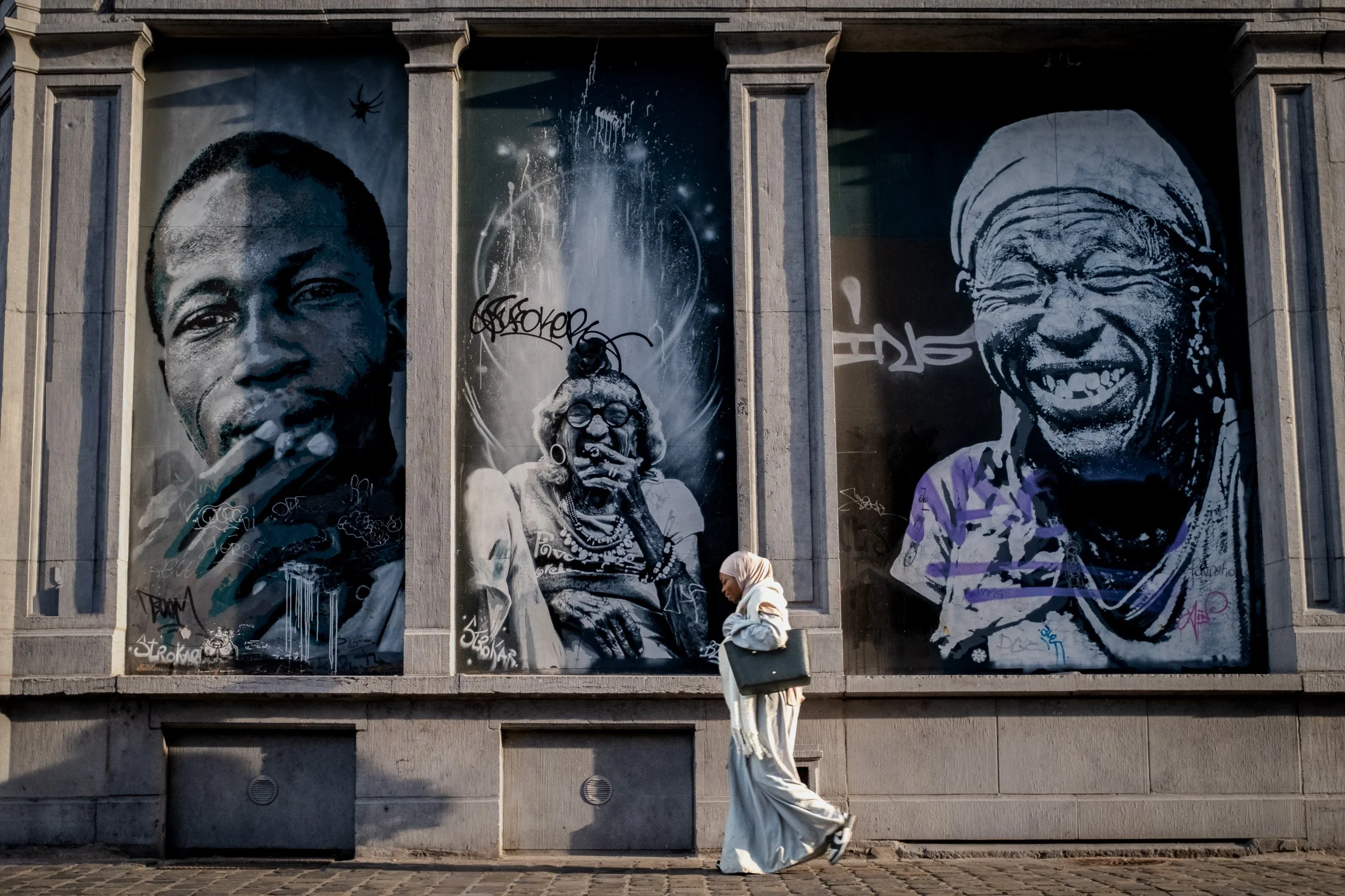 Street art mural featuring three black and white portraits of diverse women on a building wall with graffiti tags, a person walking and a purple photo on the right.