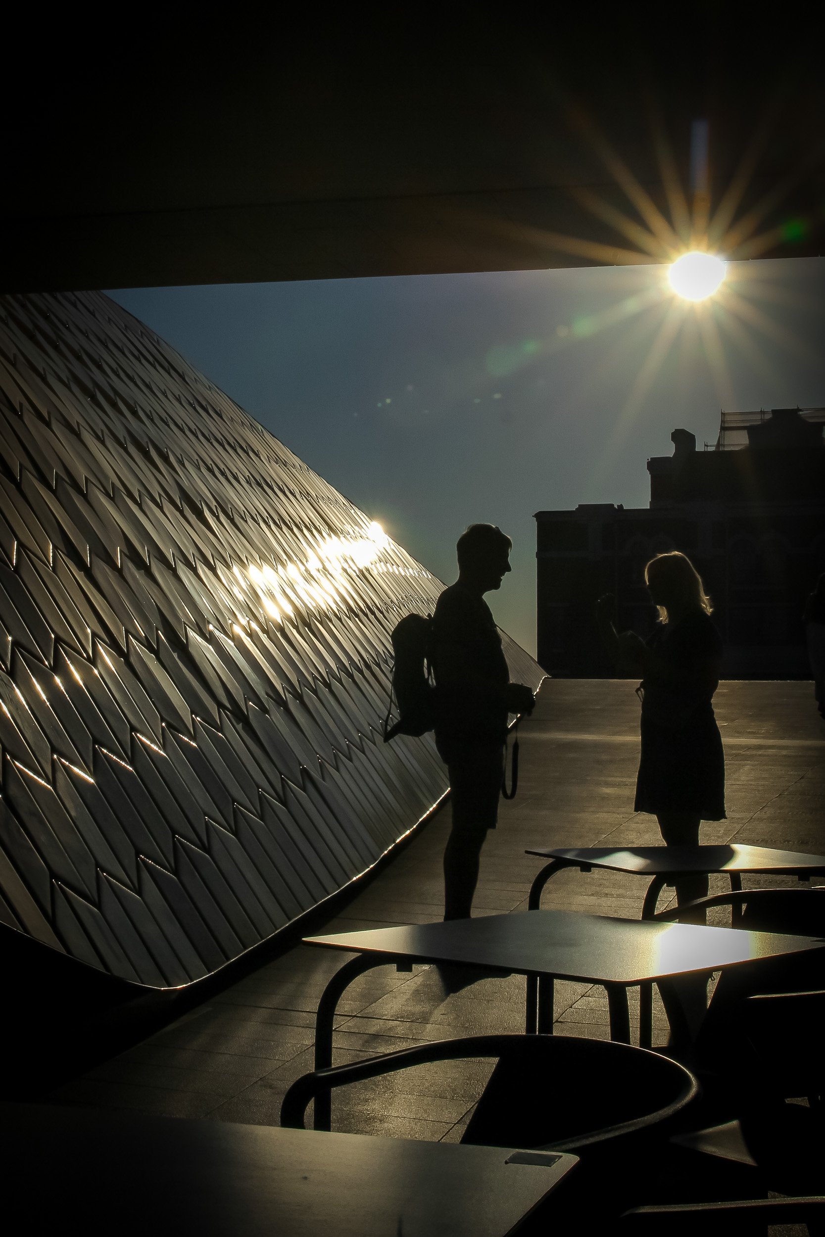 Silhouettes of two people talking outdoors during sunset, with sunlight creating starburst effect and reflections on nearby building surface.