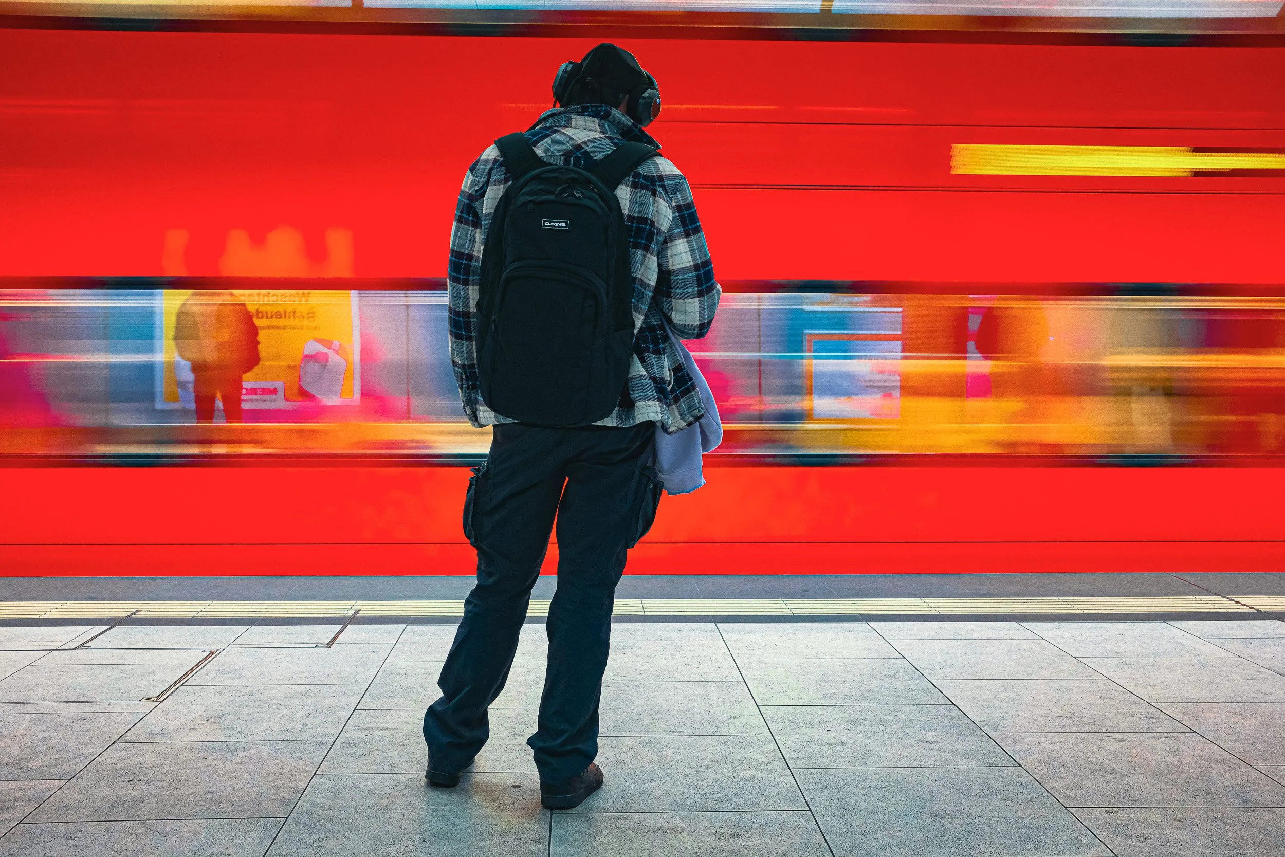 Passenger wearing headphones and a backpack stands still as a red train blurs past.