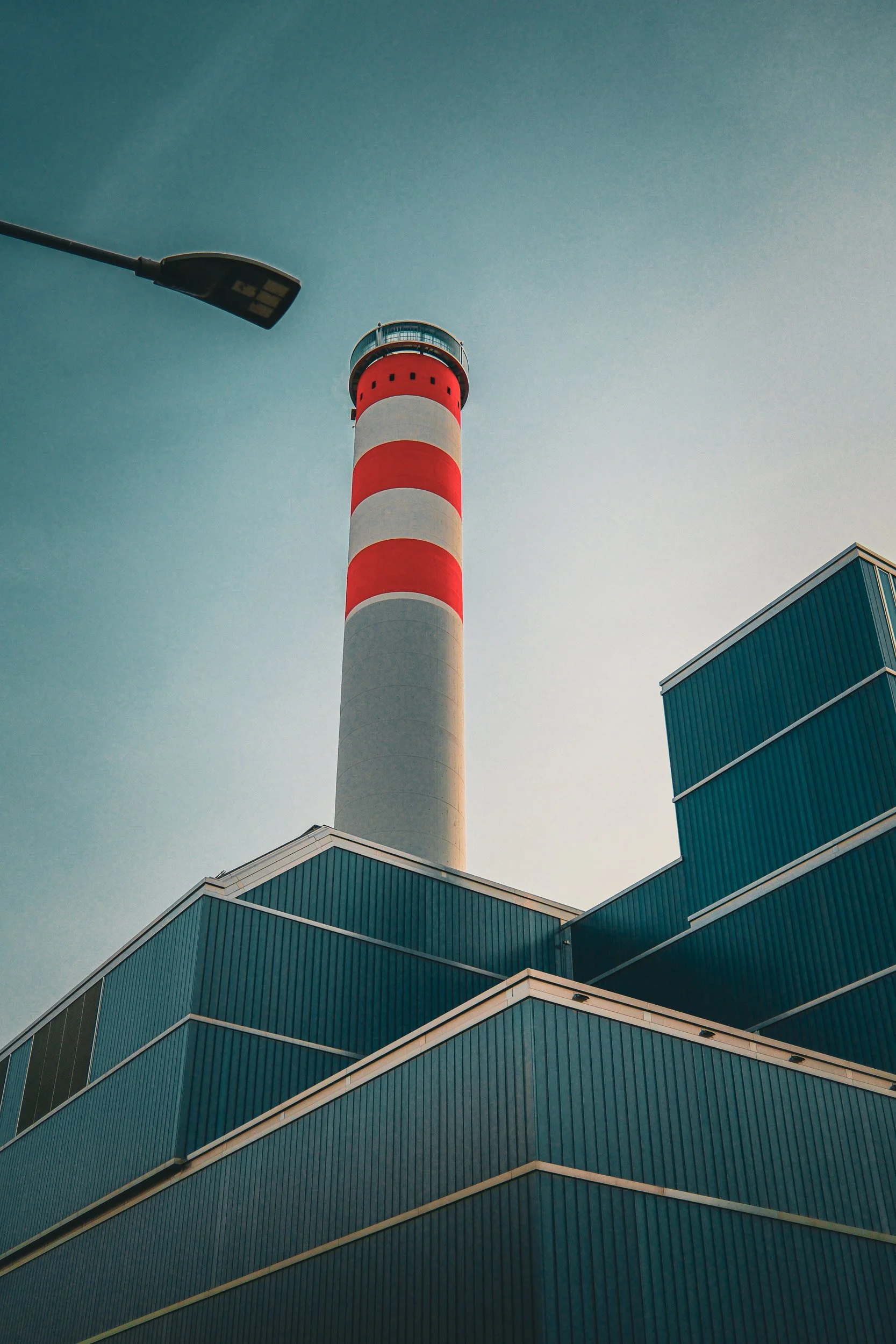 Red-and-white striped industrial chimney rises above angular blue metal roofs beside a street lamp.