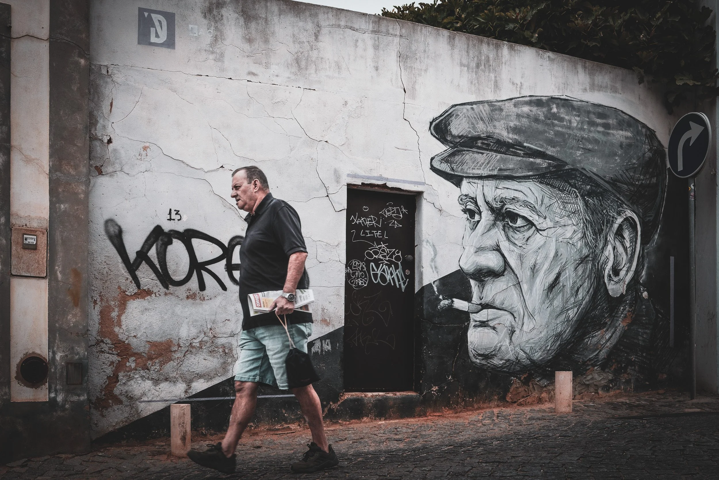 Man carrying a newspaper walks beside a mural of an older man in a cap smoking a cigarette.