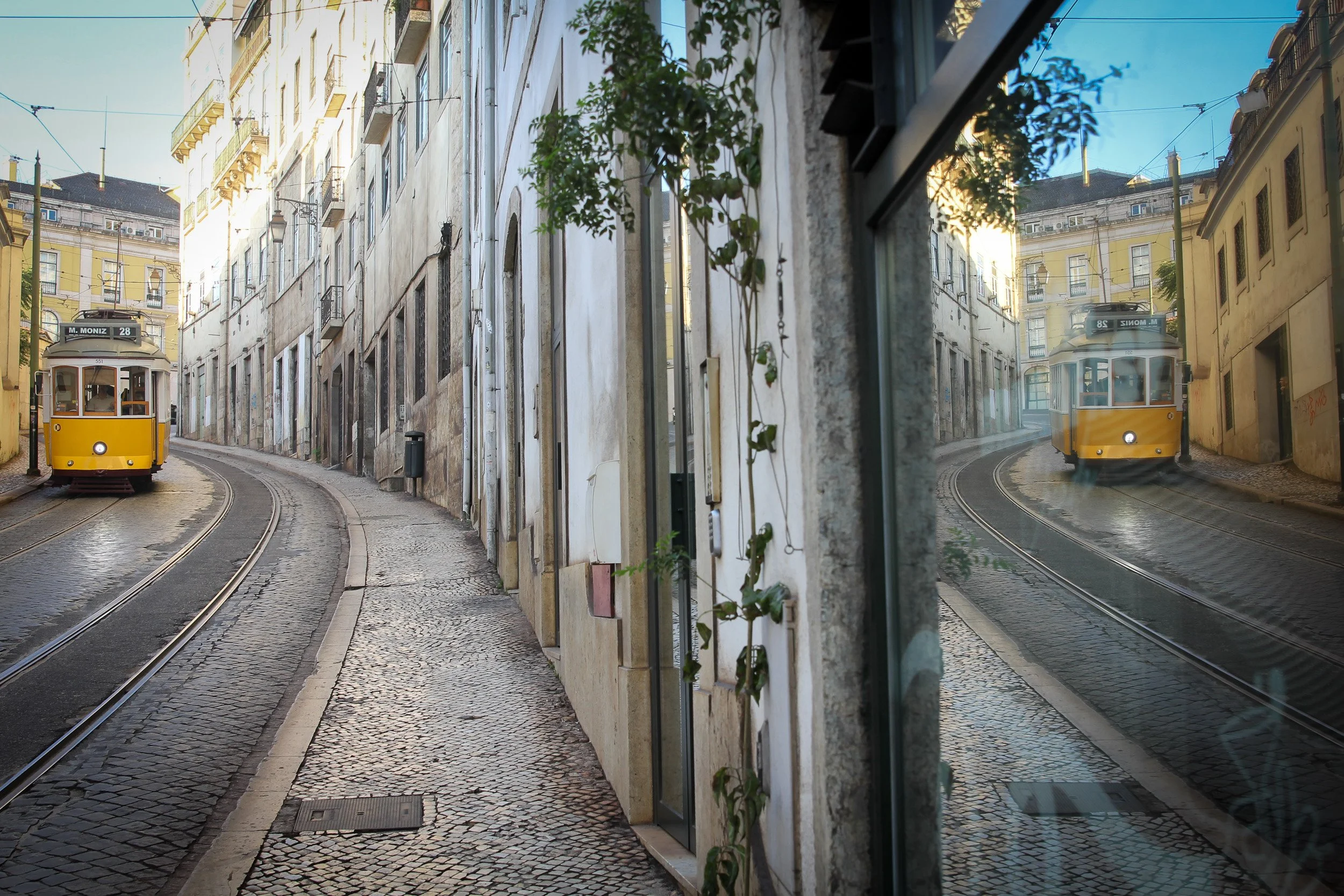 Yellow tram travels along a narrow cobbled street beside its reflection in a shop window.