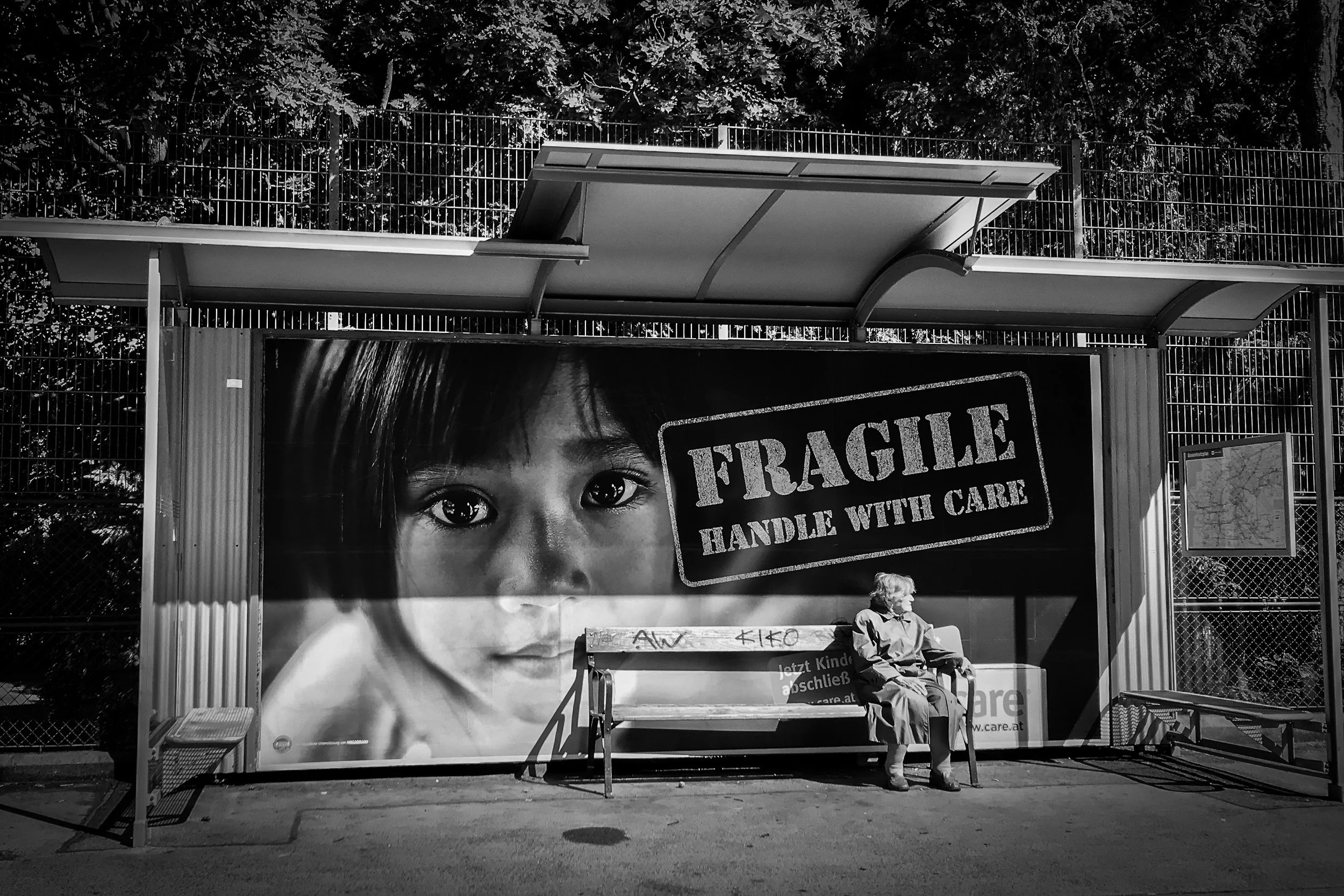 Black and white photo of a bus shelter with a large poster of a child's face and a 'Fragile' warning sign. An elderly person is sitting on a bench under the shelter.