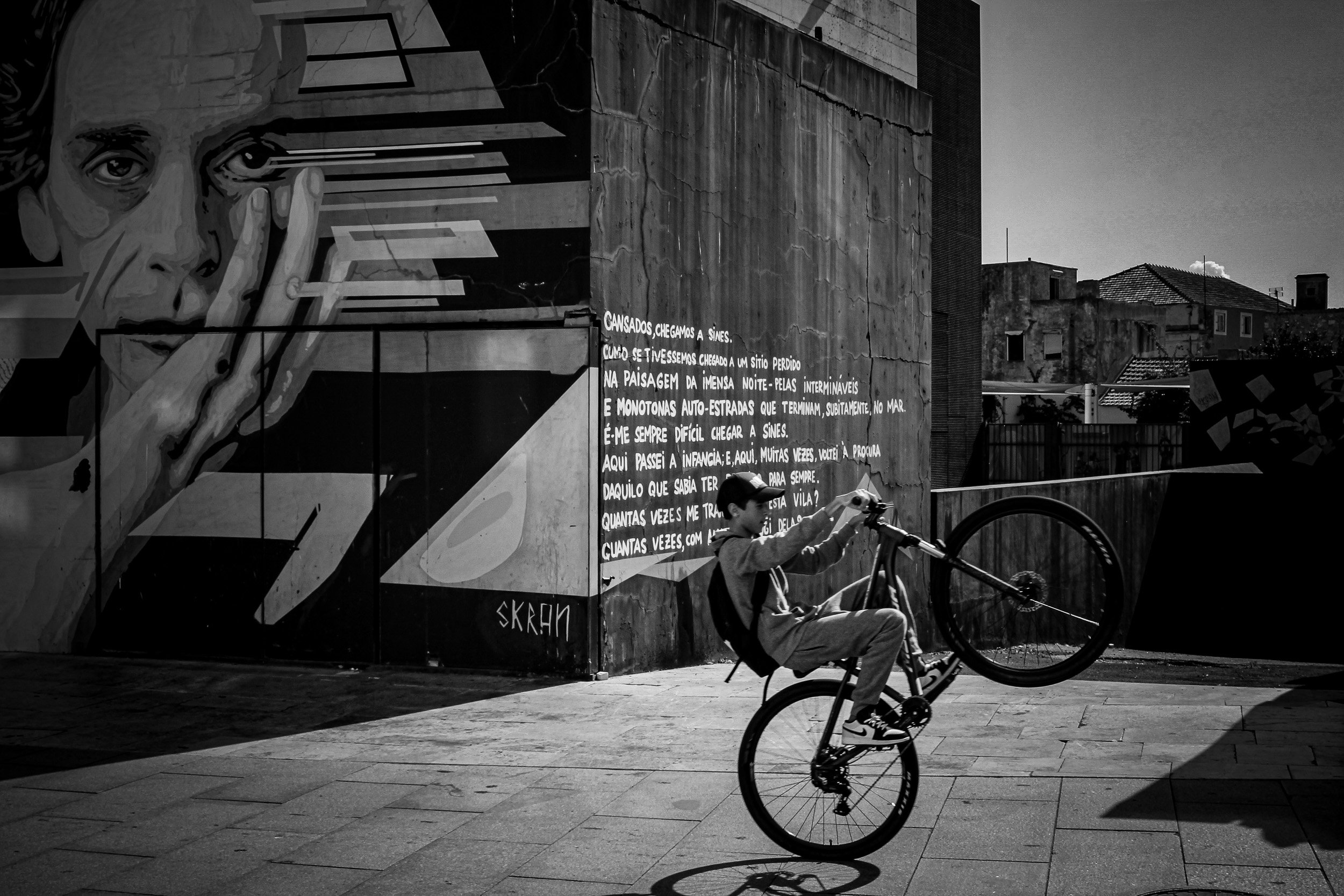Cyclist performs a wheelie in front of a large black-and-white mural portrait and handwritten text.