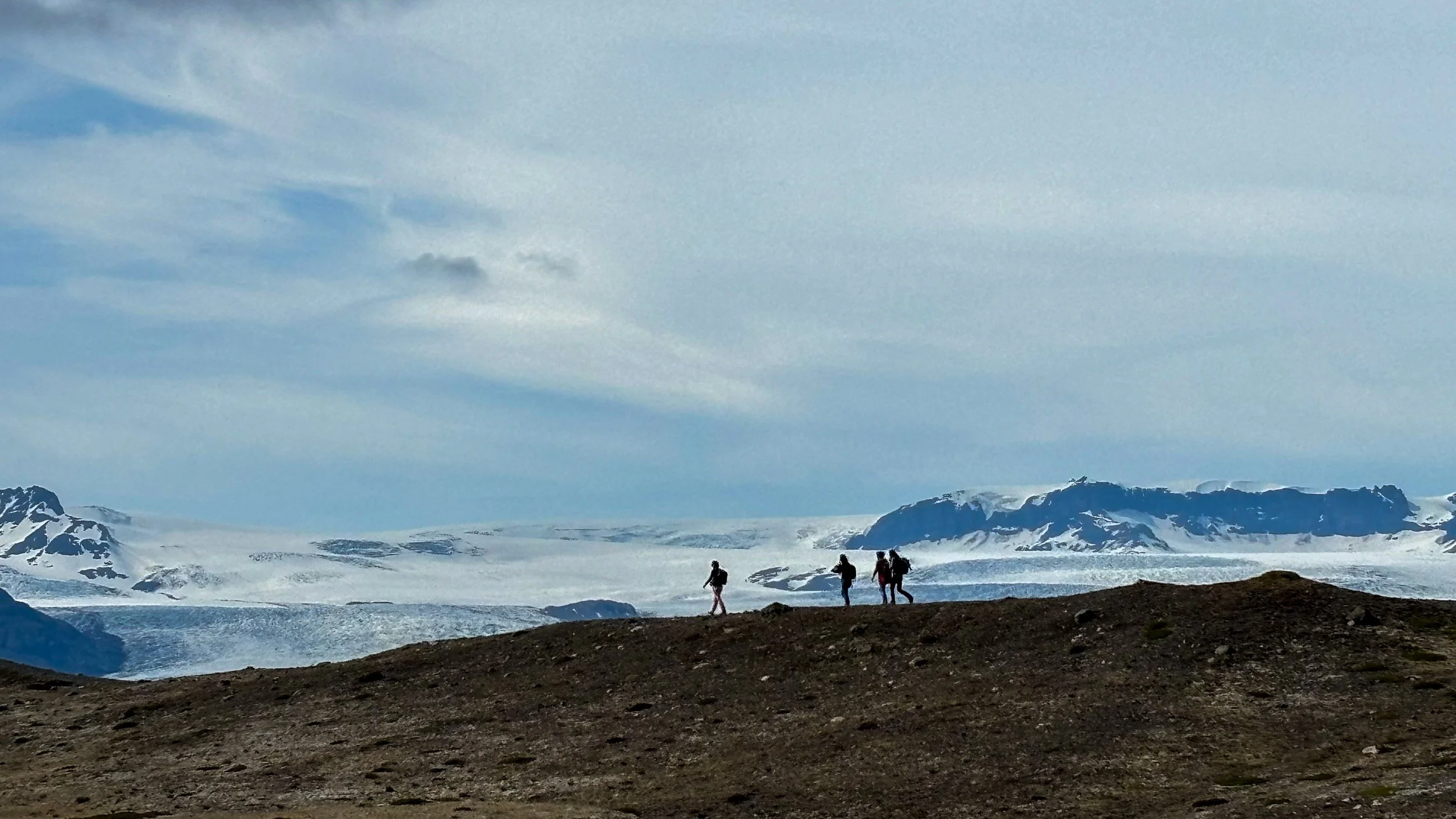 Four hikers cross a dark ridge in front of a vast glacier and snow-covered mountains.