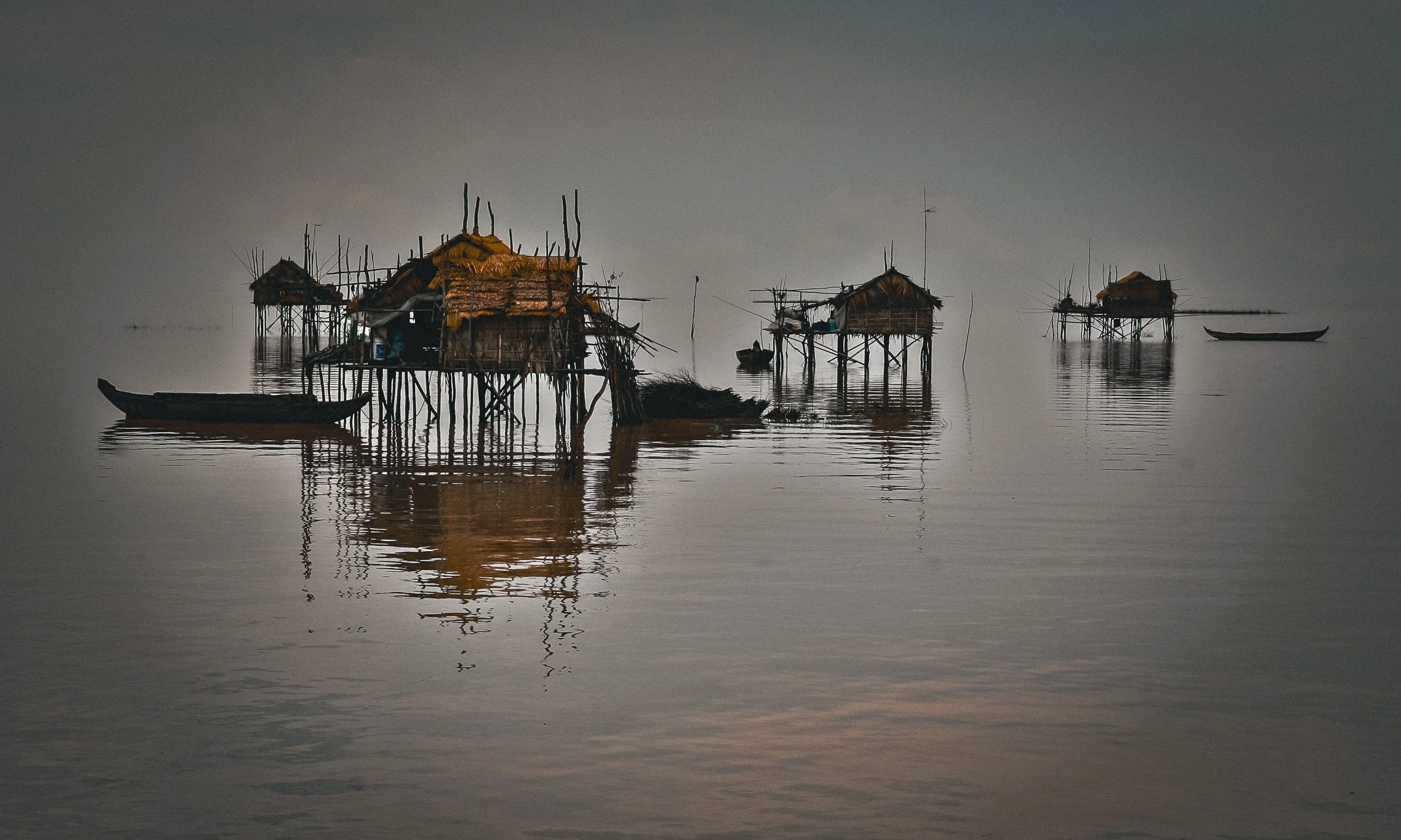 Fishing huts on stilts and small boats rest on calm water beneath a hazy sky.
