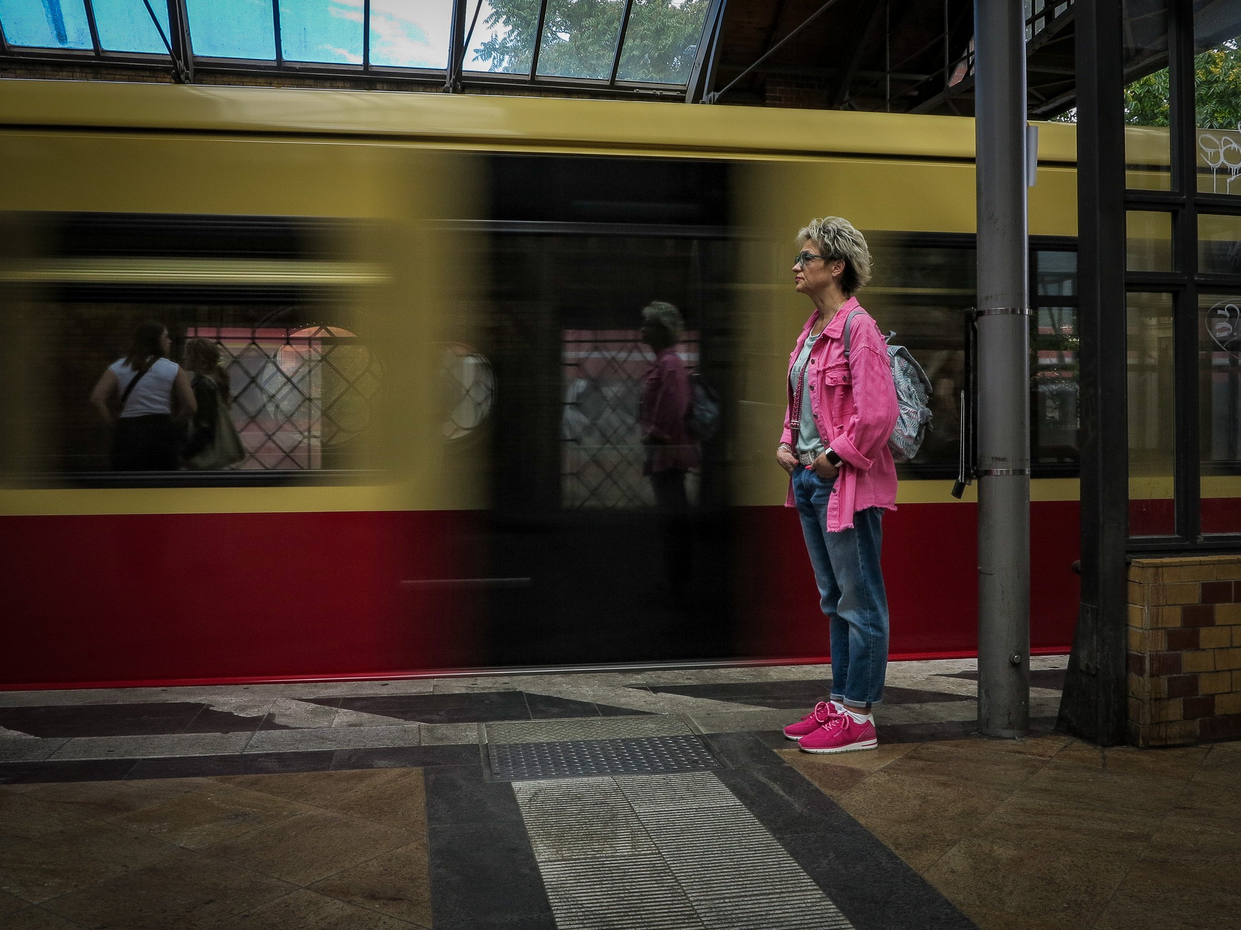 A woman with short blonde hair, glasses, pink jacket, and pink shoes standing at a bus stop, with a yellow bus passing by.
