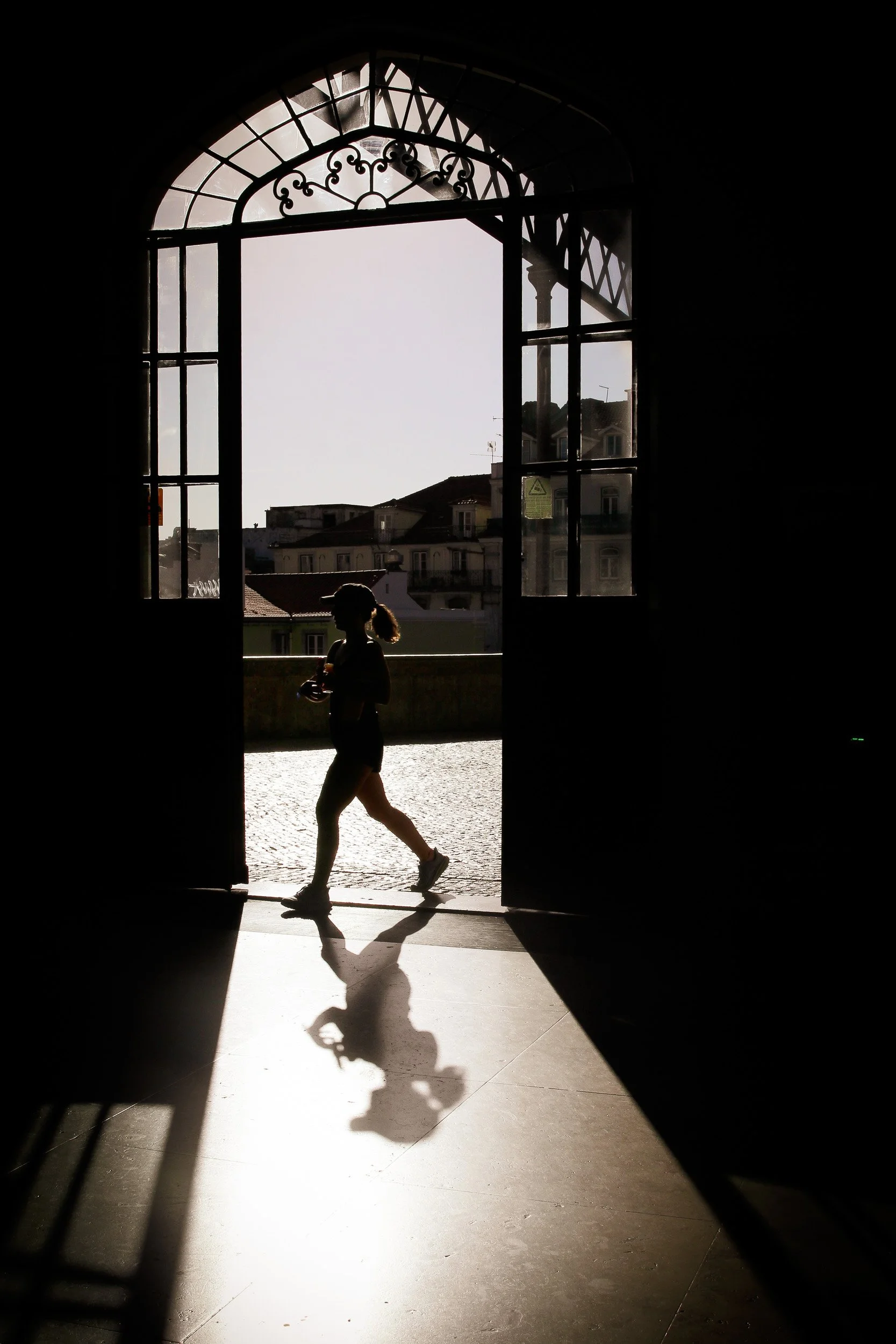 Silhouetted person walking through a sunlit arched doorway, casting a long shadow across the floor.