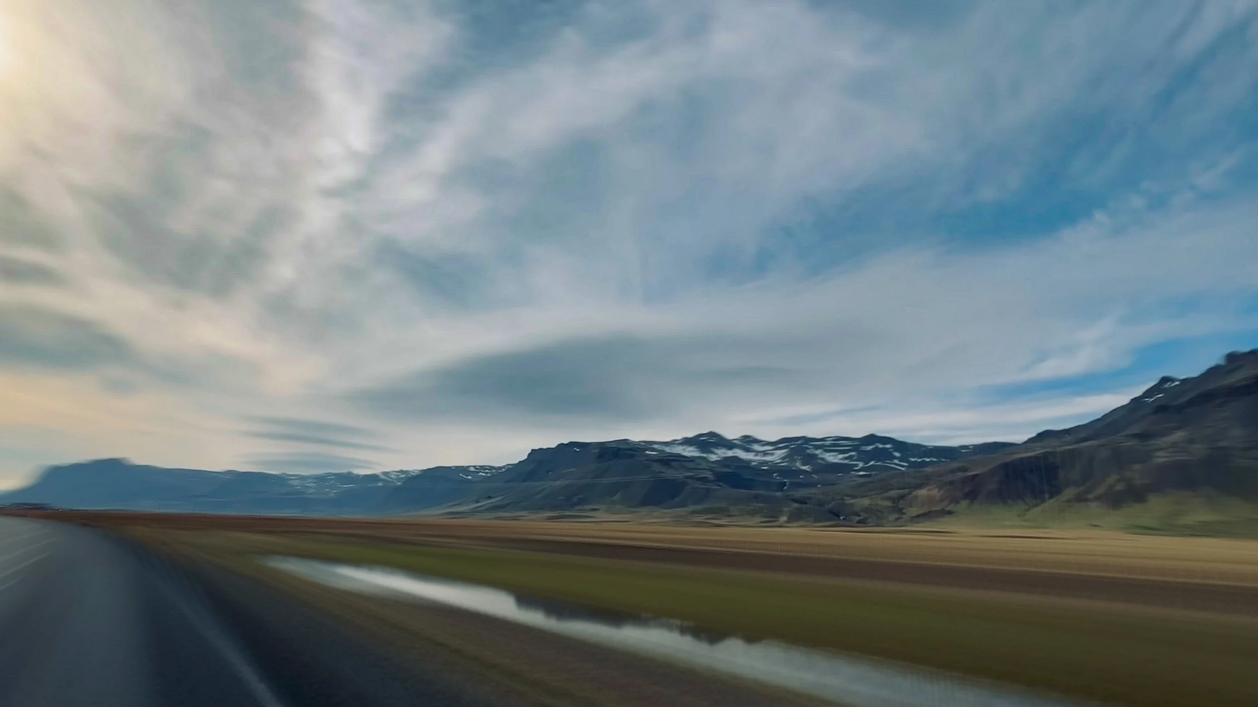Open road crosses wide plains toward snow-streaked mountains beneath an expansive sky.