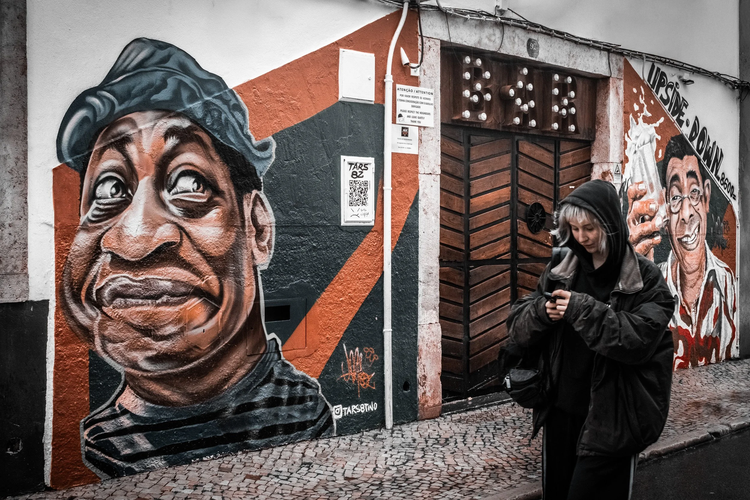 Young woman in dark clothing walks past two colorful mural portraits on a city wall.