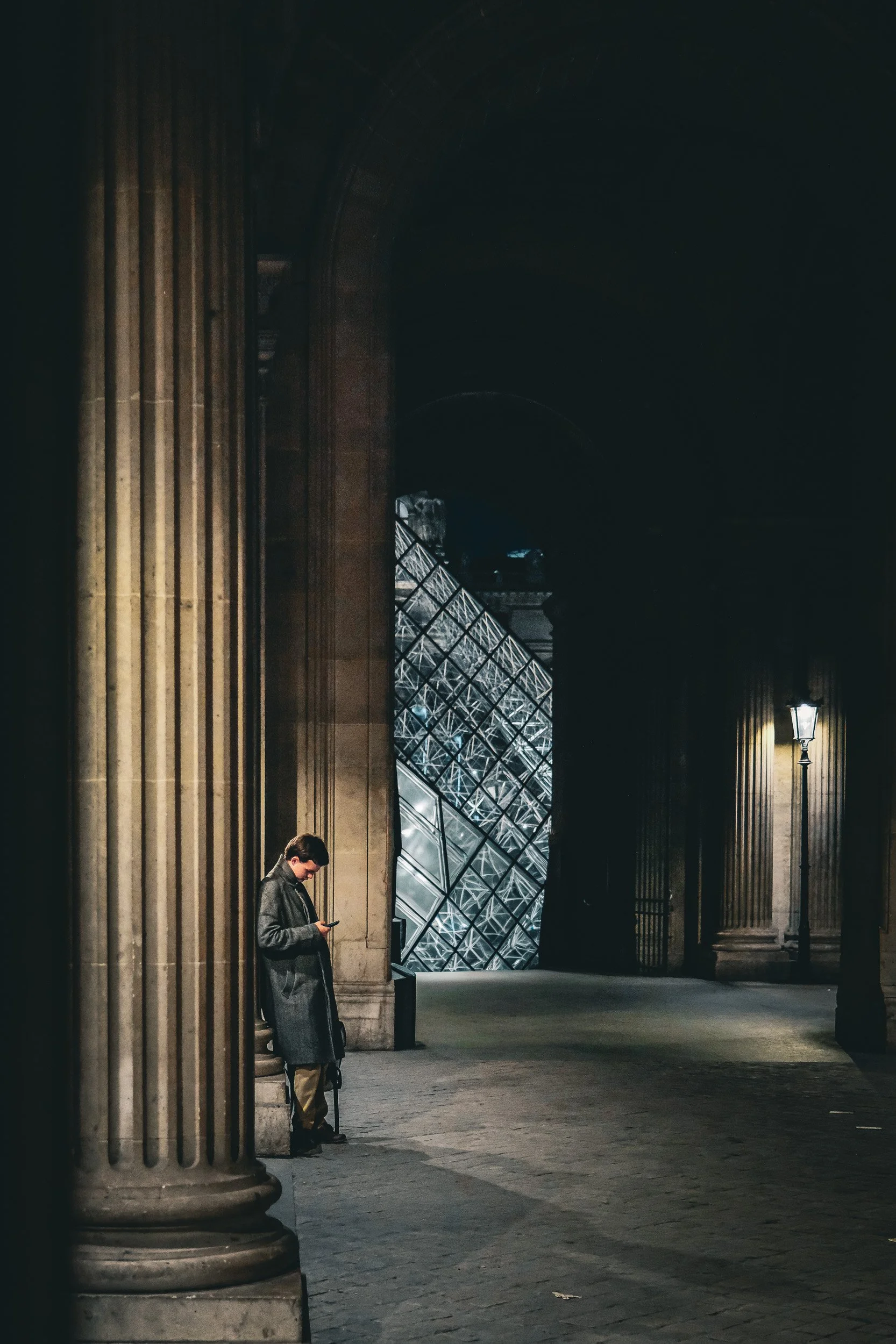 A man in a long coat standing outside a building at night, looking at his phone with one hand in his pocket, next to a large column and a lamppost, with the Louvre Museum's glass pyramid visible in the background.