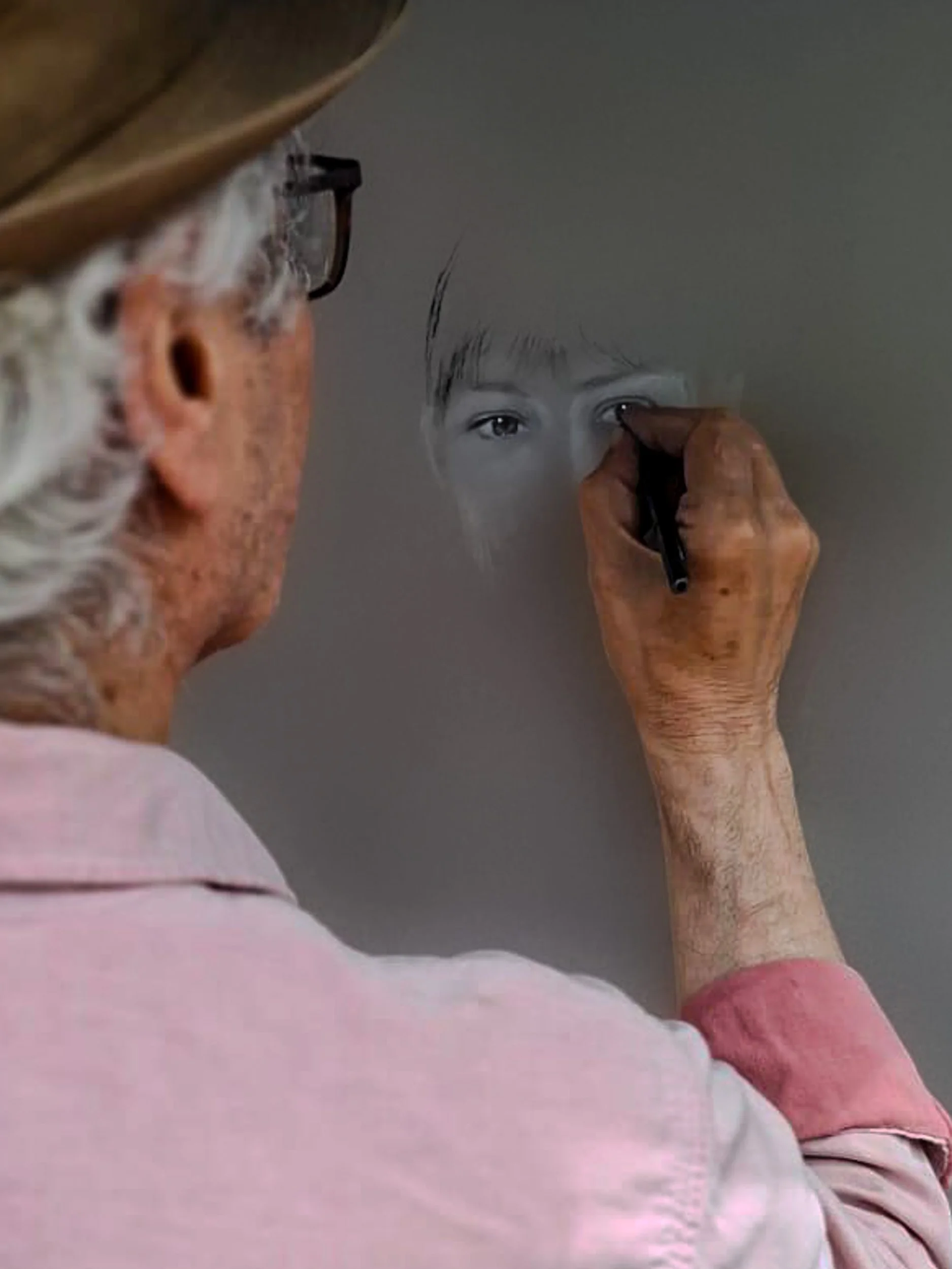 An elderly man with curly gray hair, wearing glasses and a light pink shirt, is drawing a pencil portrait of a young person on a gray surface.