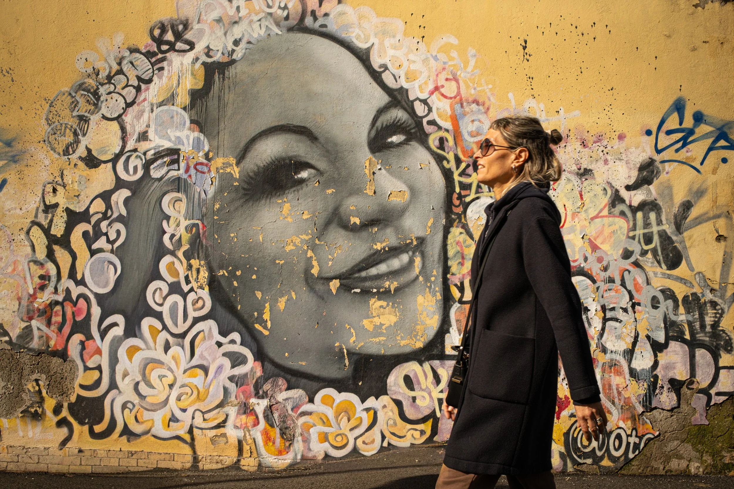 Woman in a black coat and sunglasses walks past a large smiling female mural.