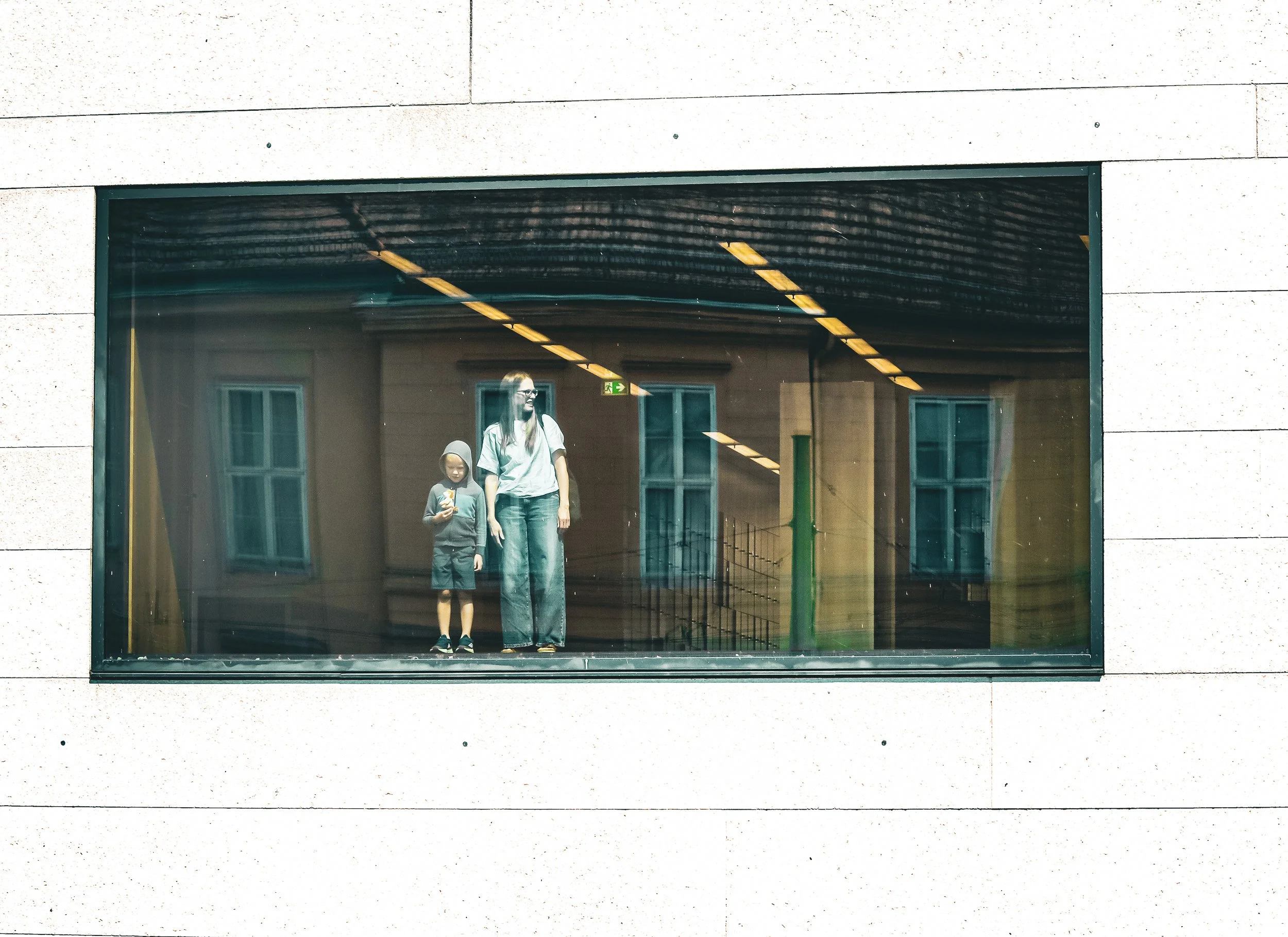 A woman and a young girl are visible through a large glass window, with the woman smiling and looking to the side while the girl holds a snack. The reflection shows a building exterior with windows and a roof structure, and indoor ceiling lights are also reflected in the glass.