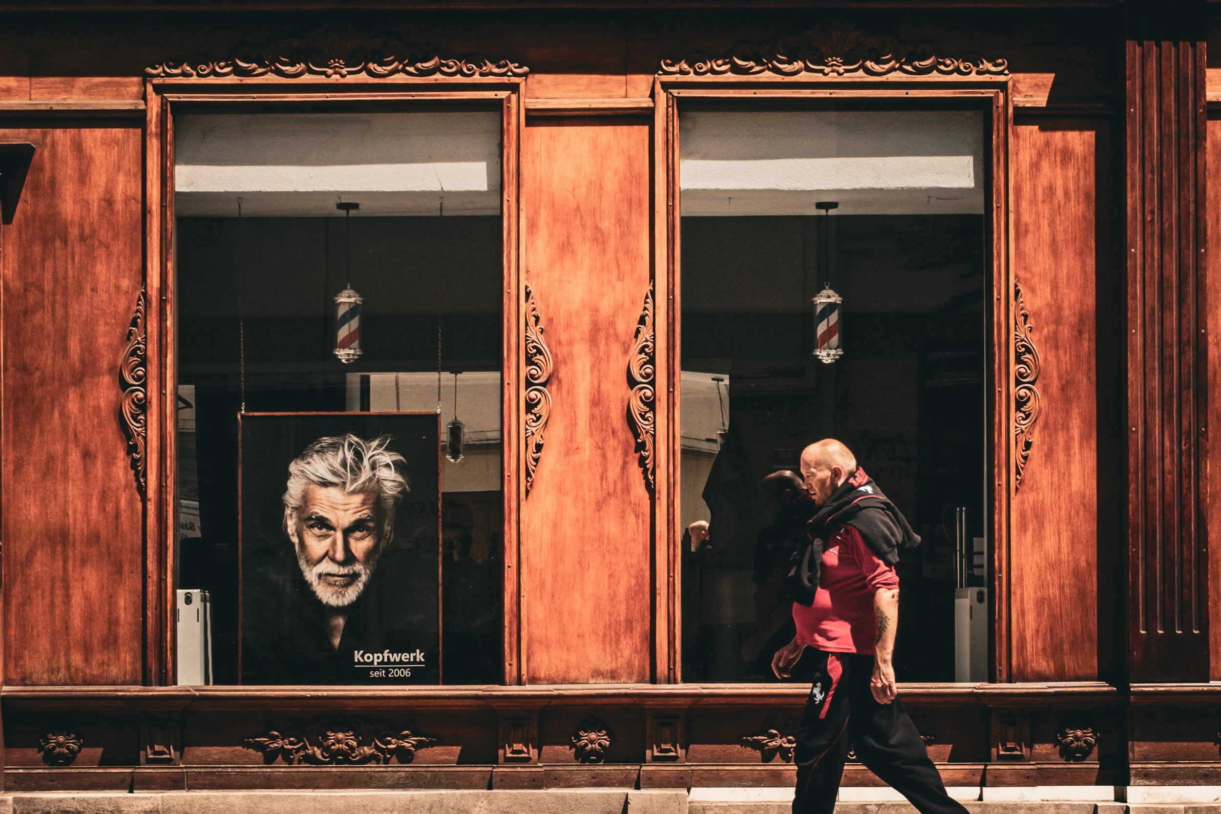 Older man in a red shirt walks past a barber shop window displaying a grey-haired portrait.
