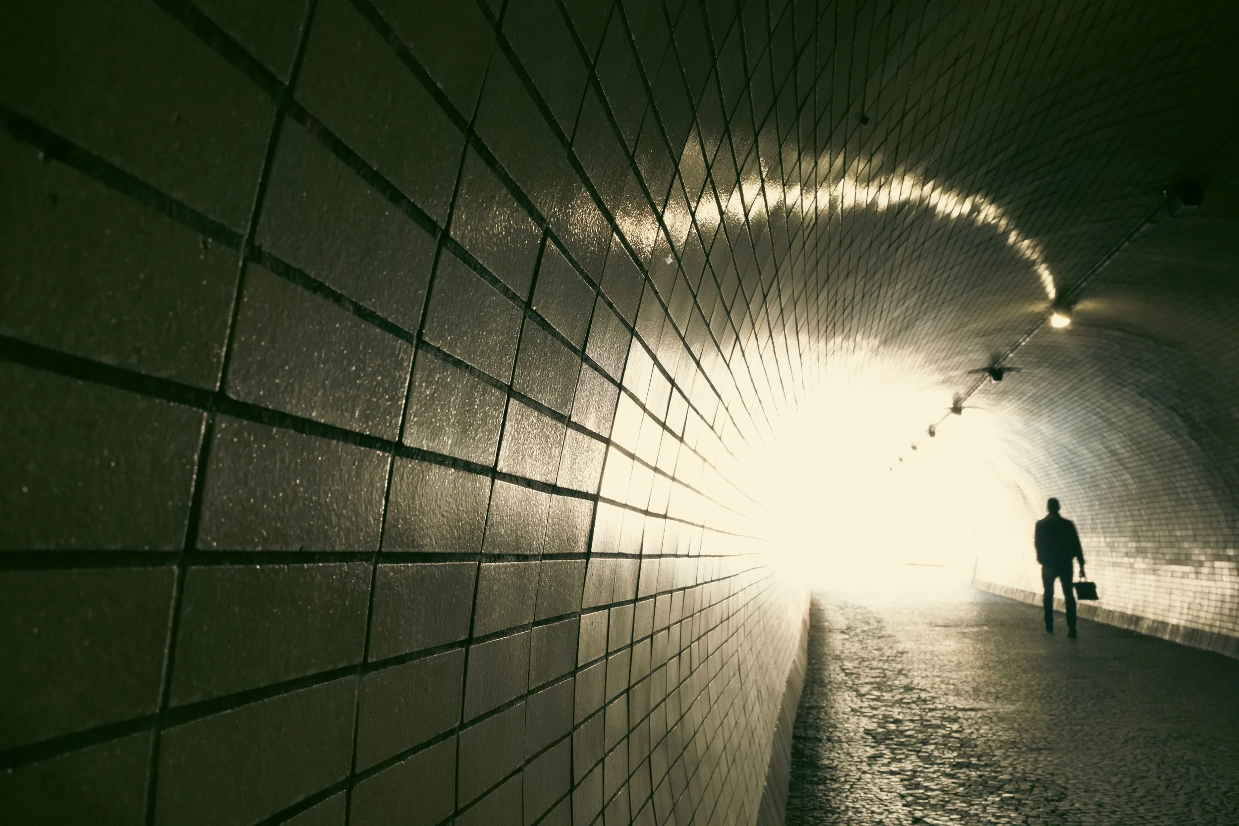 A person walking through a tunnel with brick walls and a cobblestone floor, illuminated by bright light at the other end.