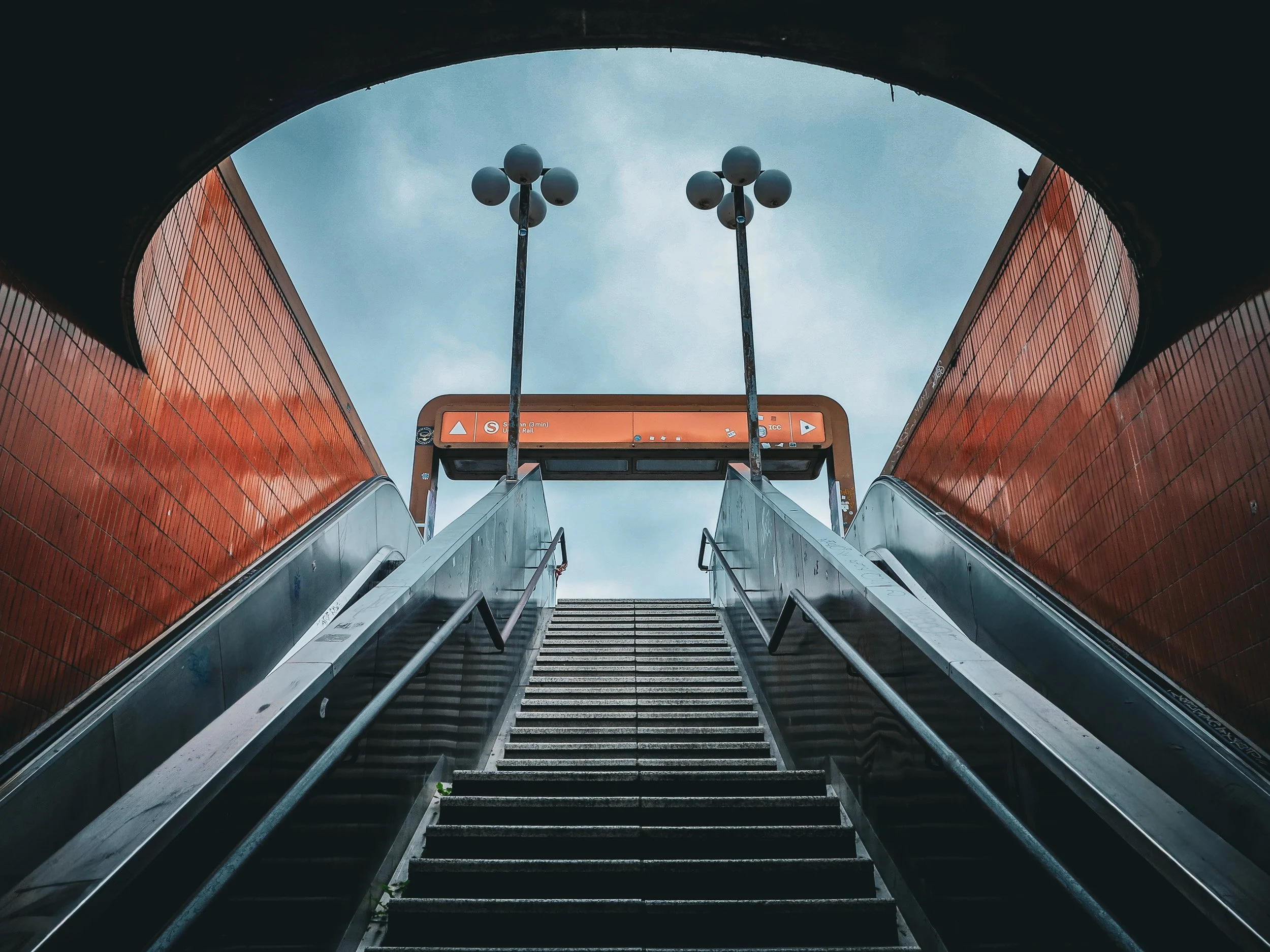 Symmetrical stairs and escalators rise between red brick walls toward twin street lamps beneath a curved opening.
