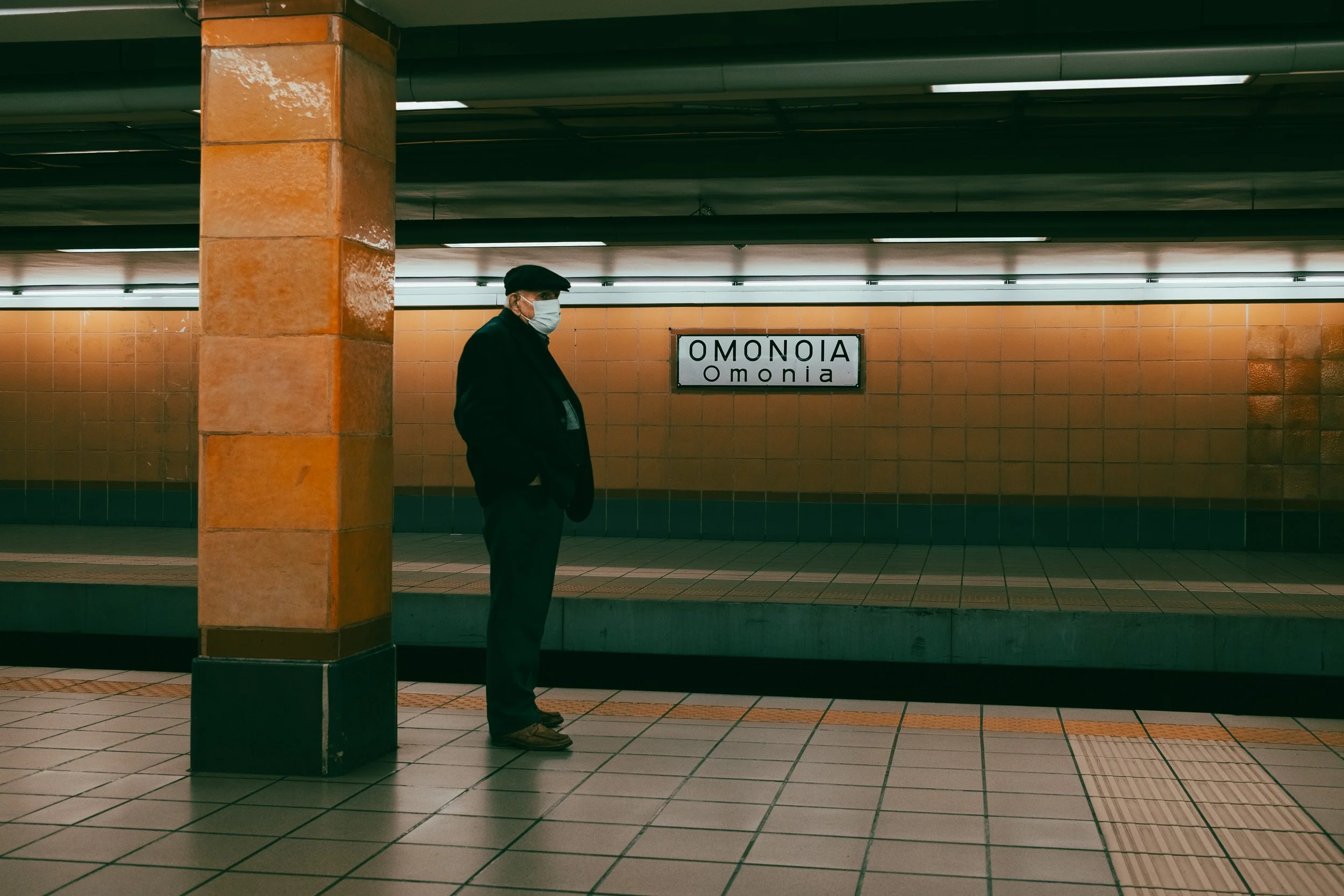 Man in a flat cap and face mask waits beside an orange column on the Omonia metro platform.