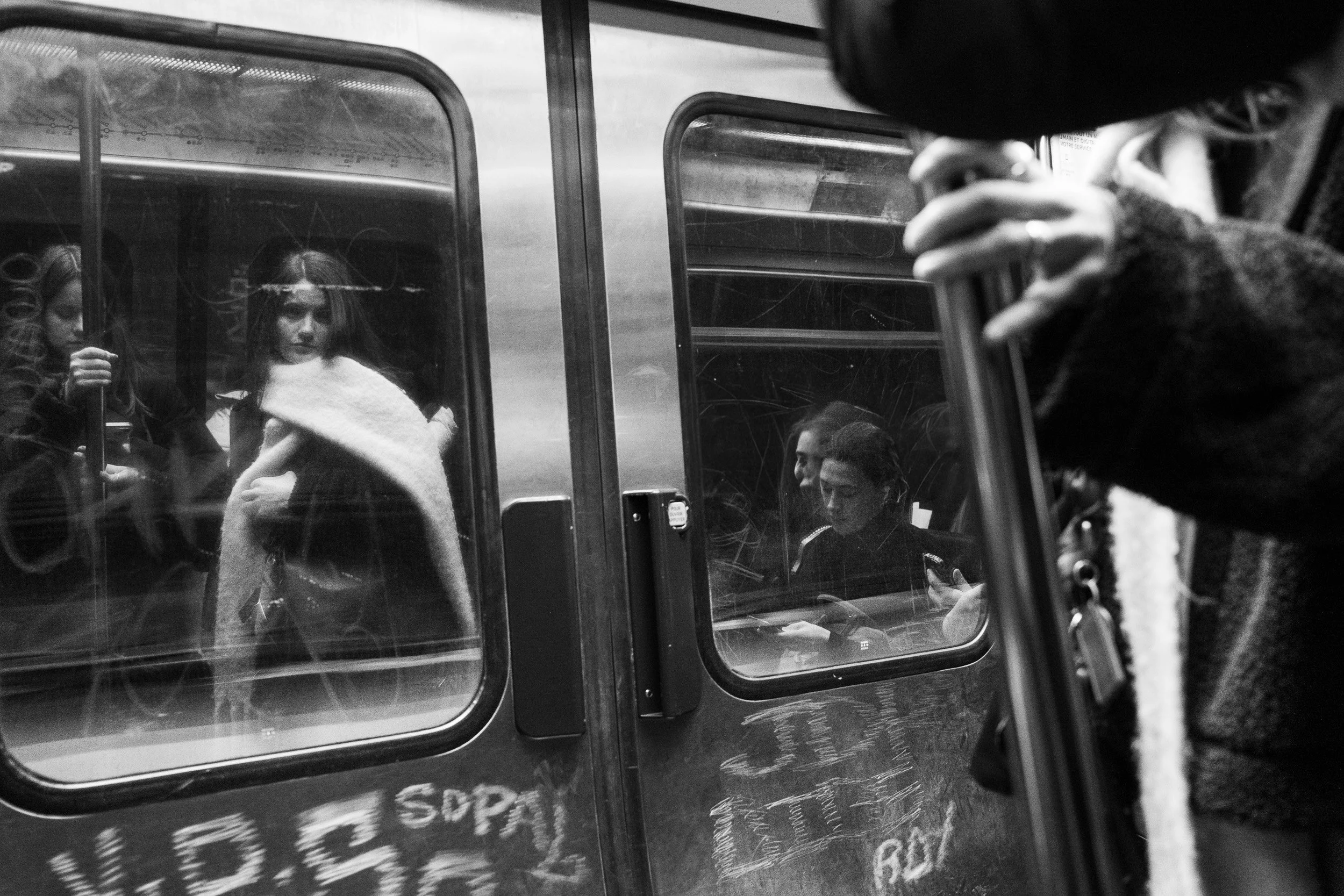 Black and white photo showing several people through subway train windows. Some appear to be waiting or sitting, with one person looking at their phone and others with headphones or holding drinks. The scene captures a busy urban environment.