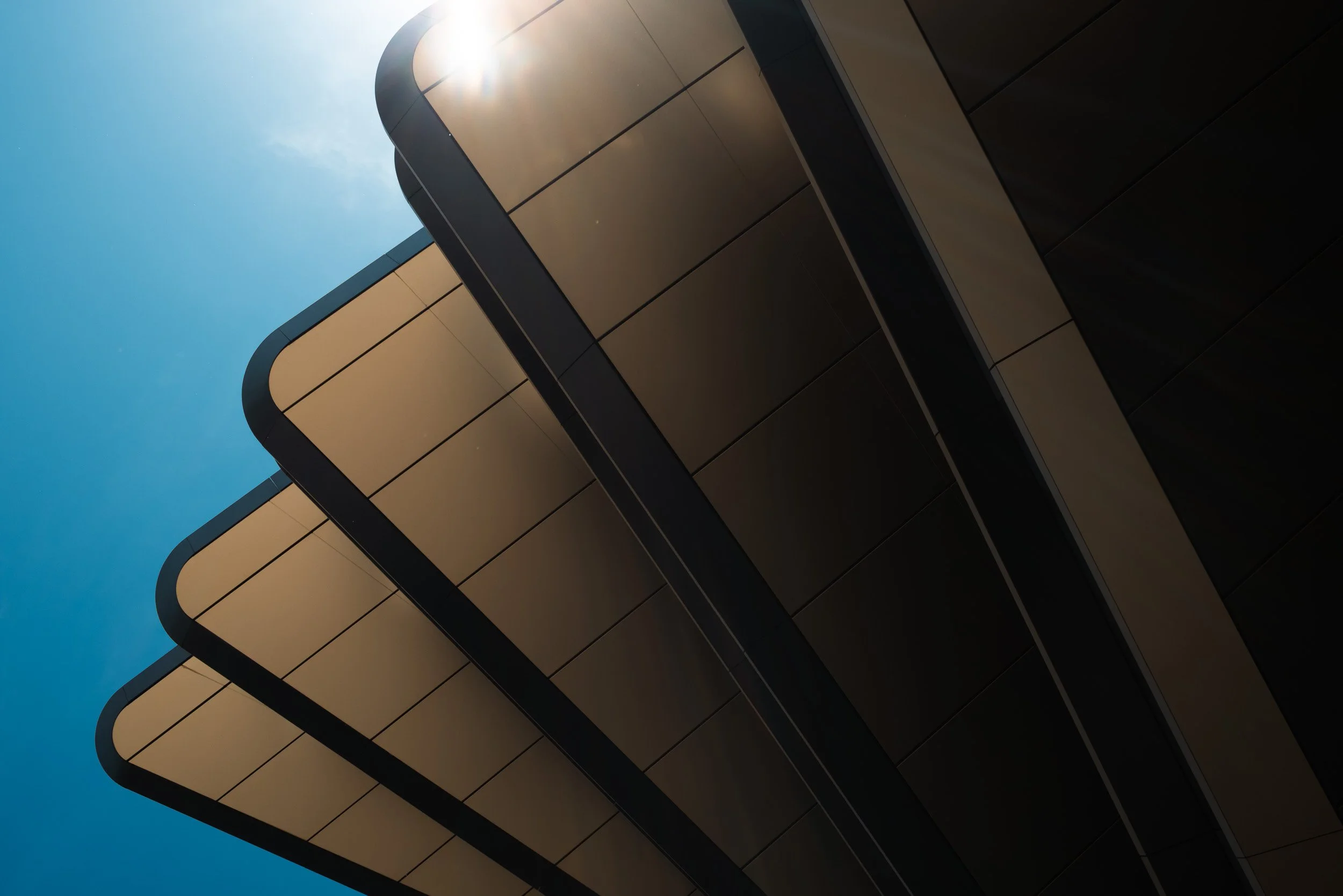Rounded modern building overhangs recede against a deep blue sky with sunlight at the edge.