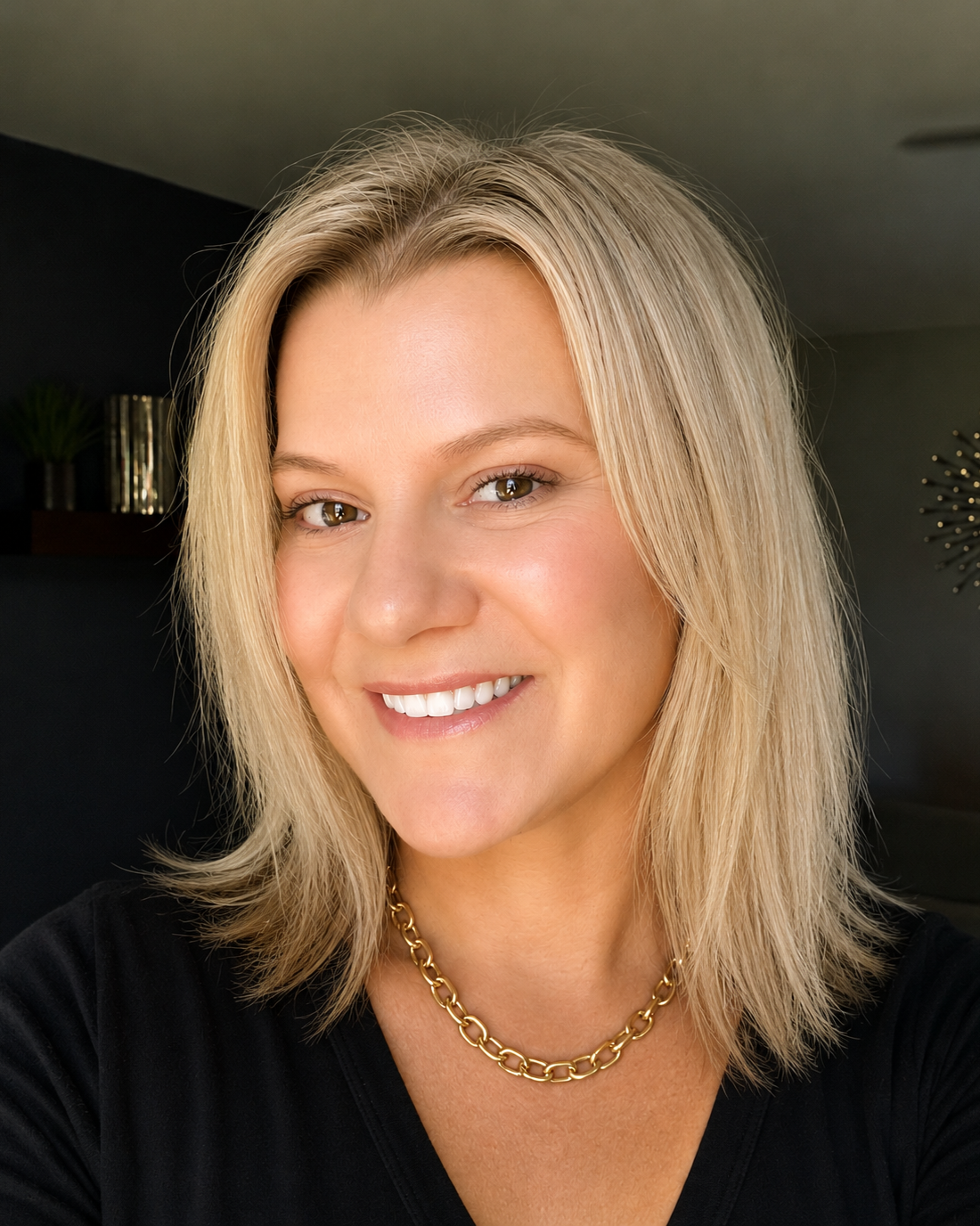 Close-up portrait of a woman with blonde hair and brown eyes smiling, wearing a black top and a gold chain necklace, in a modern indoor setting.