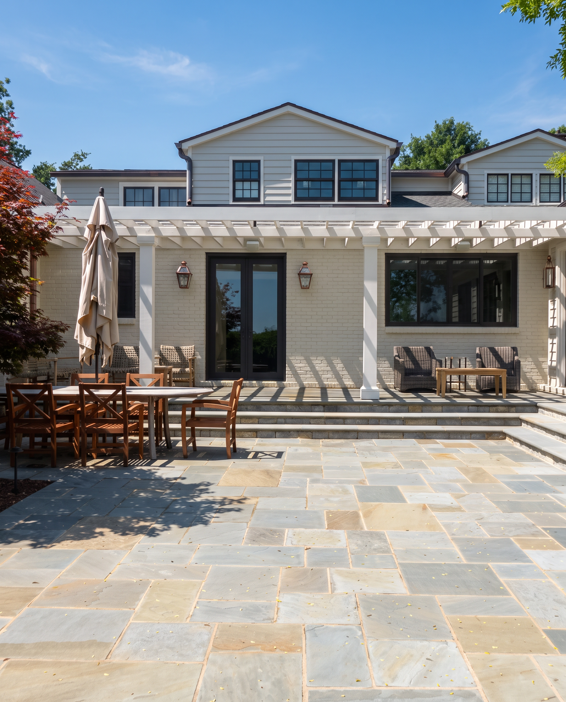 Backyard patio with stone flooring, a wooden dining table with chairs, an outdoor umbrella, and a two-story white house with black windows and a pergola structure.