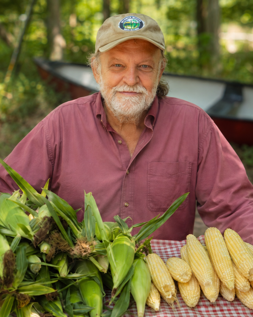 A smiling man wearing a beige cap and maroon shirt sits behind a table with fresh corn on the cob and corn still in the husks, outdoors in a wooded area.