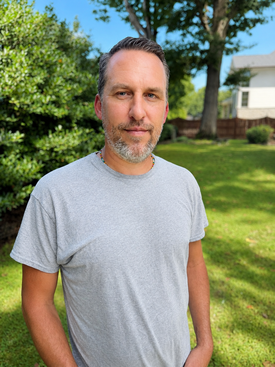 A man with short gray hair and a beard standing in a lush green backyard with trees, bushes, and a house in the background