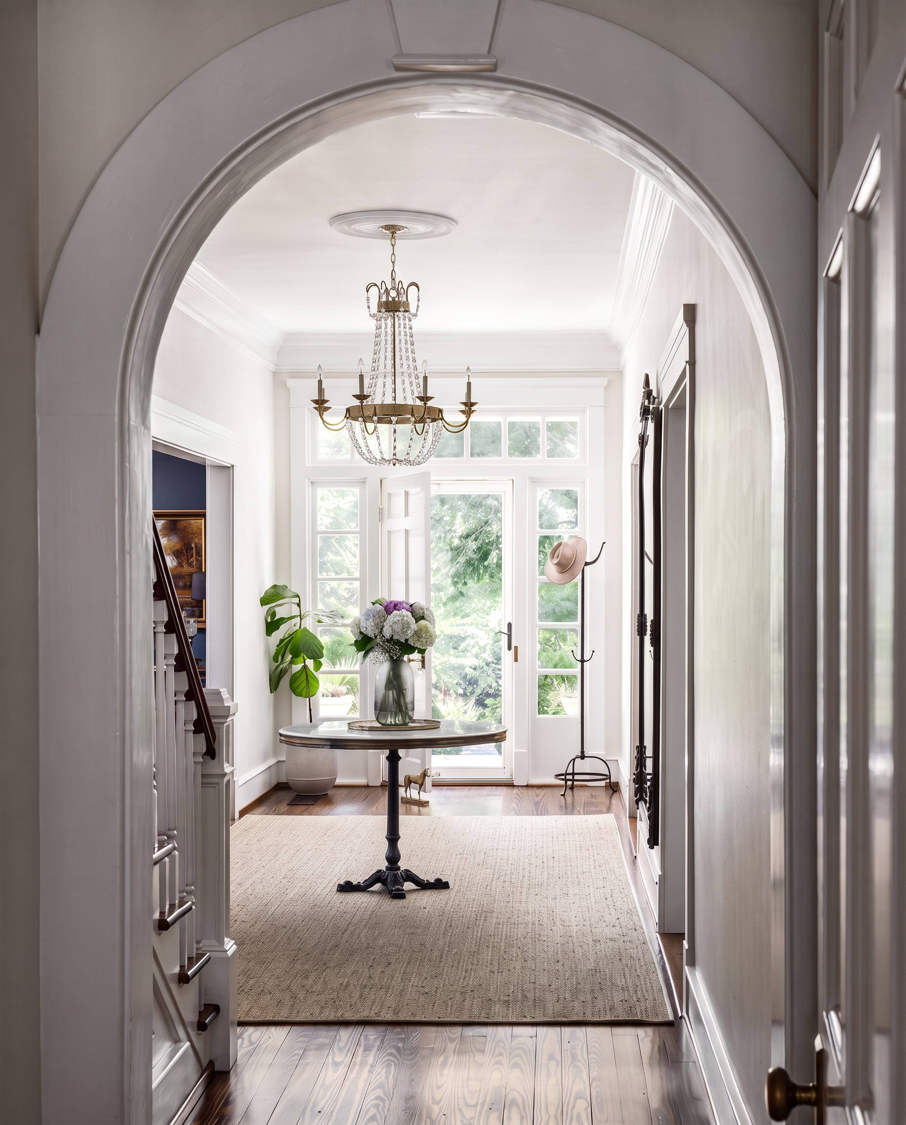 View through a white-framed doorway into a bright entryway with a chandelier hanging from the ceiling, a round table with a floral arrangement, large windows, a hat hanging on a coat rack, and a small decorative horse figure on the floor.