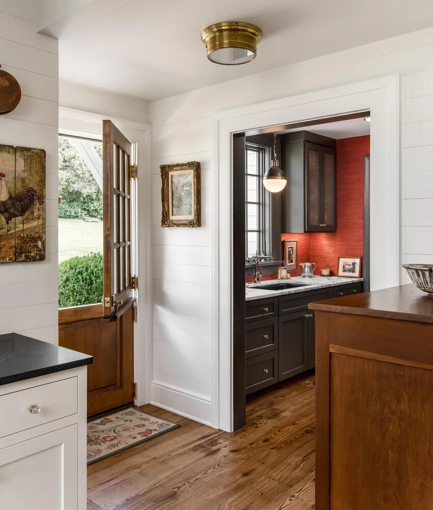 View of a cozy home interior showing a wooden front door with a window and greenery outside, leading into a living space with white walls, wooden floors, and framed artwork. An adjoining kitchen with dark cabinets, a window, and a red accent wall is 