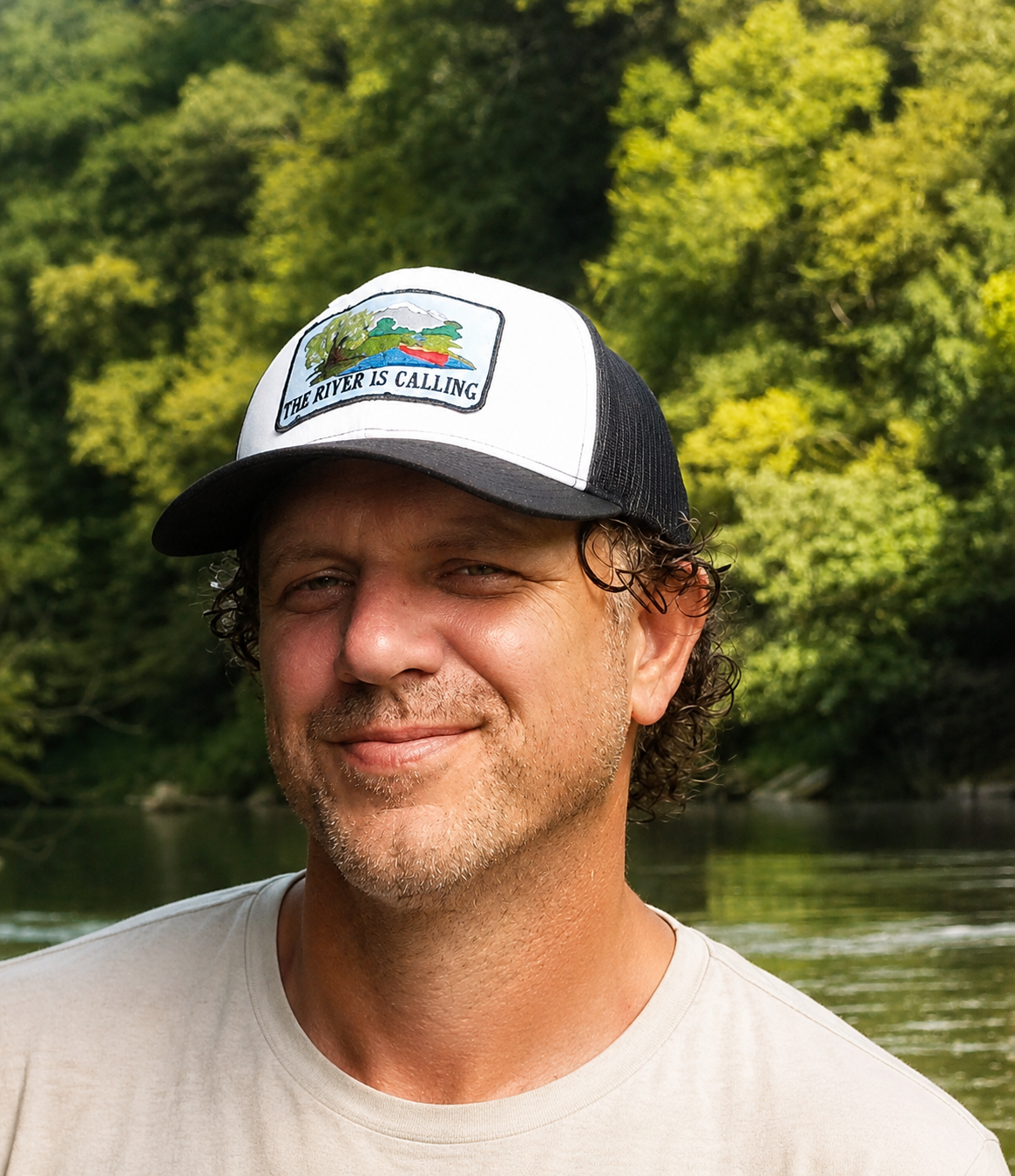 A man wearing a black and white trucker hat with a river and trees embroidered on the front, standing outdoors with a river and lush green trees in the background.