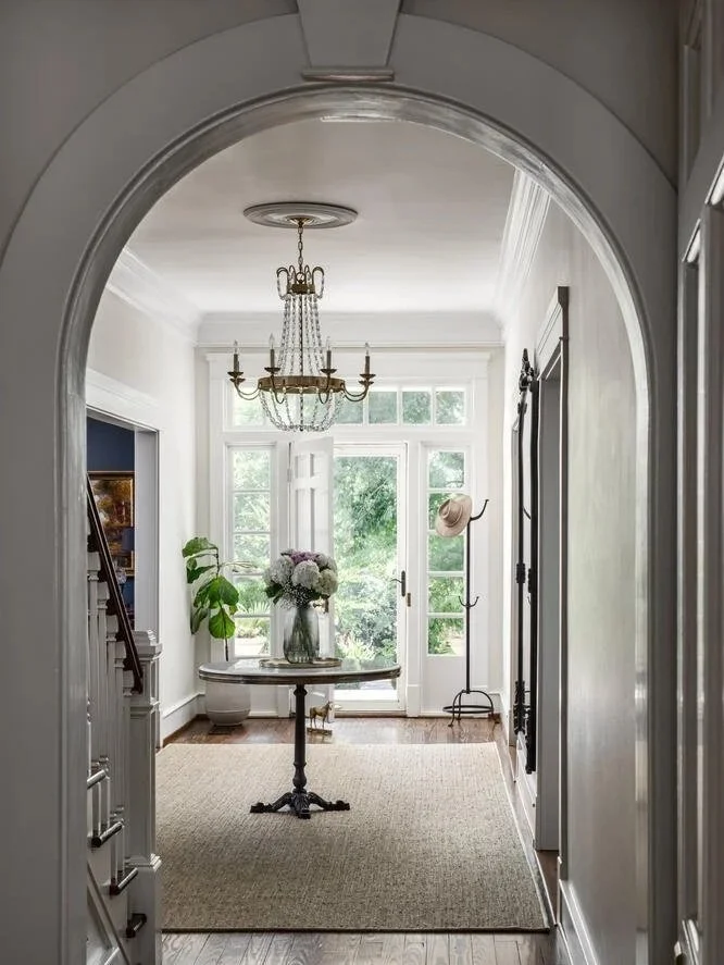 Bright entryway with a windowed door opening to greenery outside, a chandelier hanging from the ceiling, a round table with a floral arrangement, a potted plant, and a hat and coat hook on the wall.