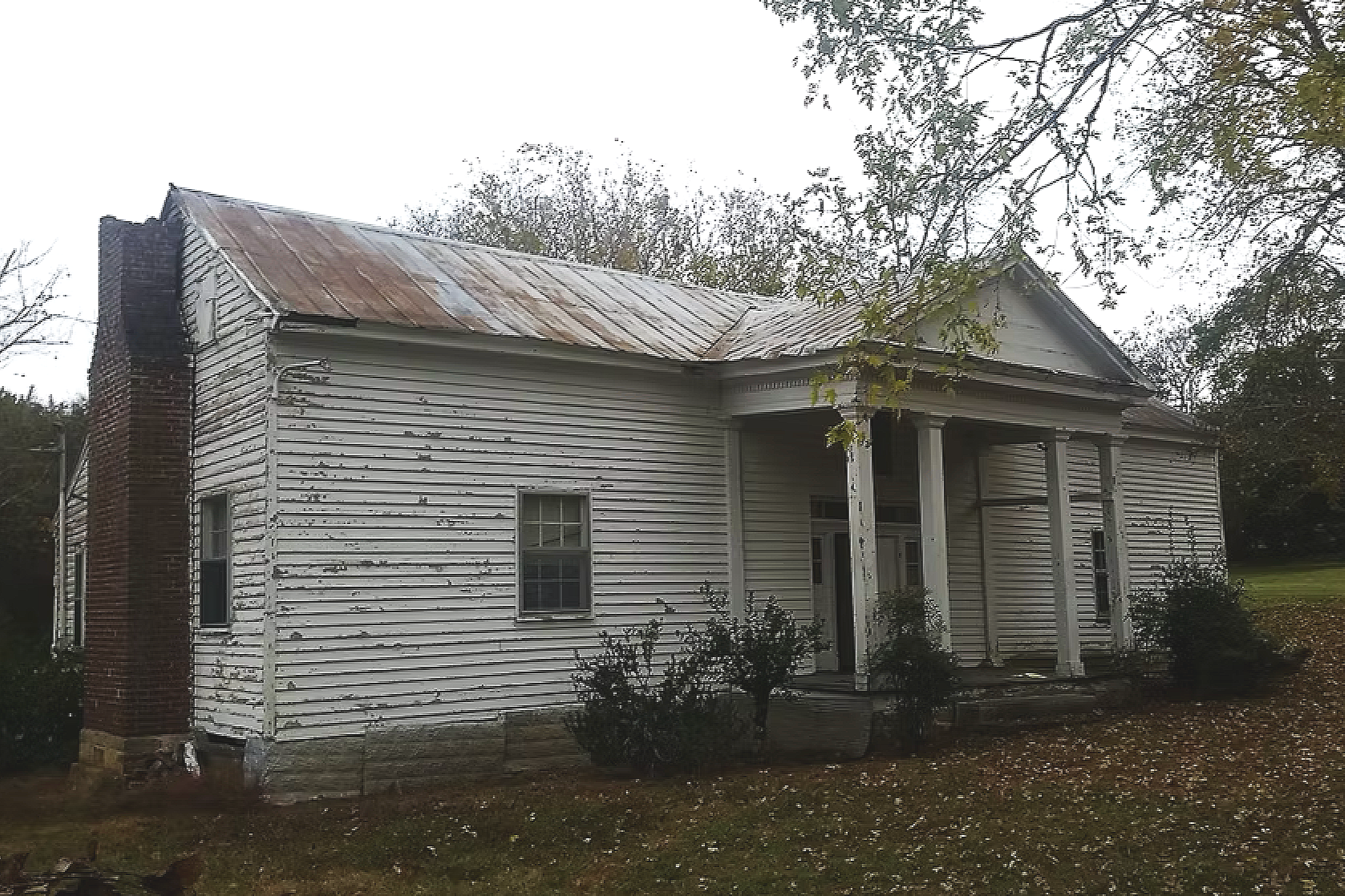 Old white wooden house with peeling paint and a rusted metal roof, surrounded by sparse trees and shrubs, under an overcast sky.