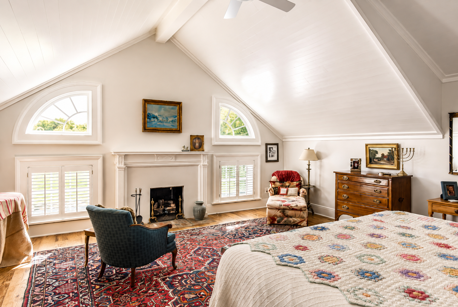 A cozy bedroom with a sloped white ceiling, white walls, and large windows with white shutters. There is a fireplace with a painting above it, and a mix of vintage furniture including a blue armchair, a red armchair, and a wooden dresser. A patterned quilt covers the bed, which is partially visible in the foreground, and a colorful rug covers the wooden floor.