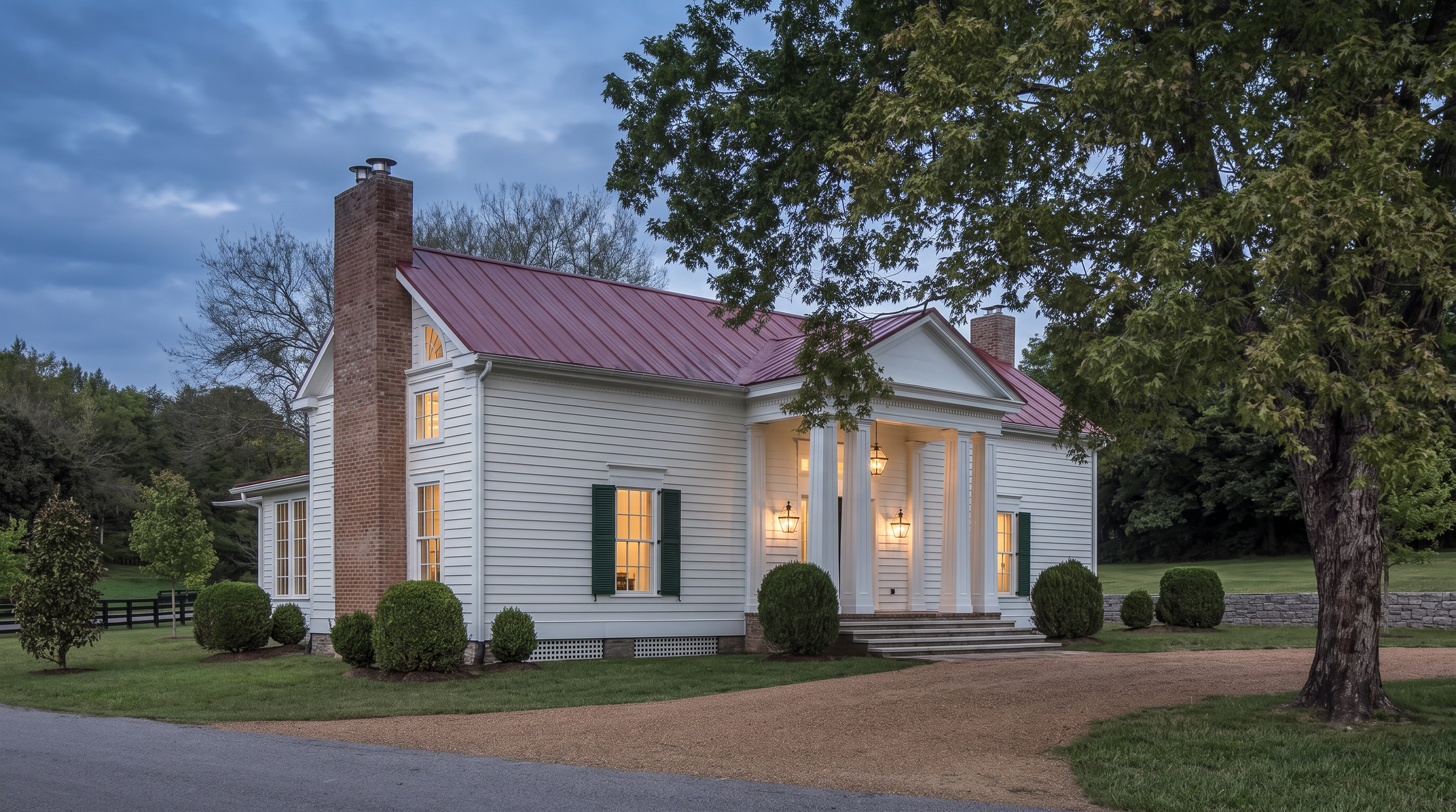 A white two-story house with green shutters and a red metal roof, surrounded by a well-maintained lawn with small bushes. There are lit lamps at the front entrance, and large trees border the property under a cloudy sky during dusk.